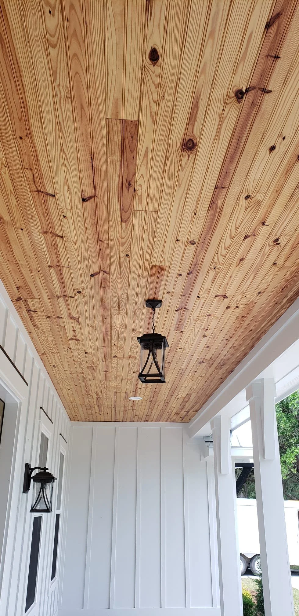 Wood-paneled ceiling with hanging lantern-style light fixtures in a porch or patio area with white walls and large window.