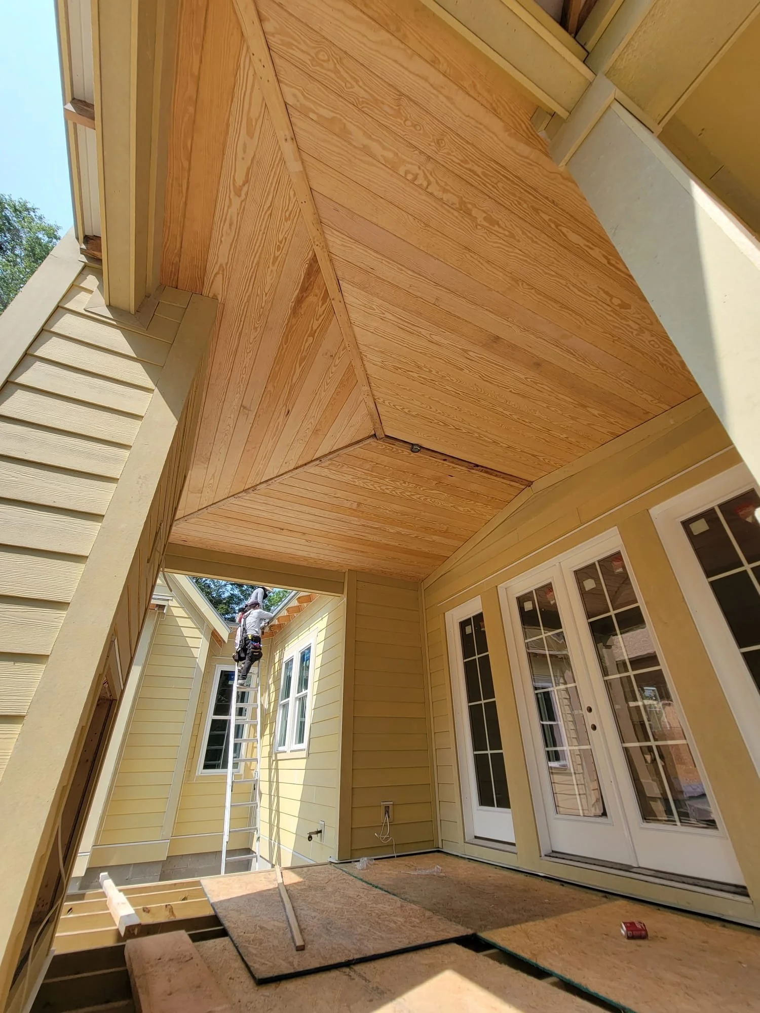 Construction worker on ladder installing a wooden ceiling in a partially enclosed porch or patio, with yellow house walls, multiple windows, and French doors.