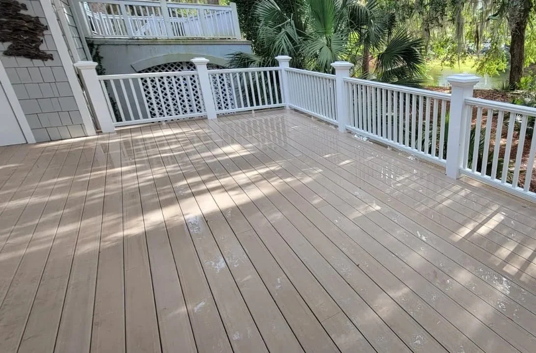 Photo of a clean, beige wooden deck with white railing, surrounded by green trees and plants, with sunlight creating shadows on the deck.