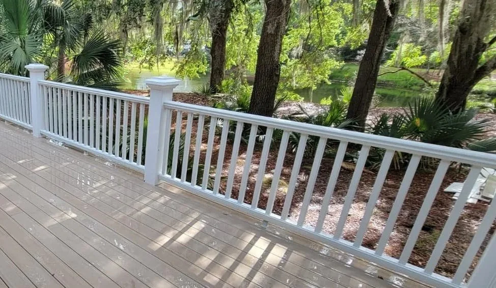 A white railing fence along a wooden deck overlooking a lush, green wooded area with trees and a small body of water.