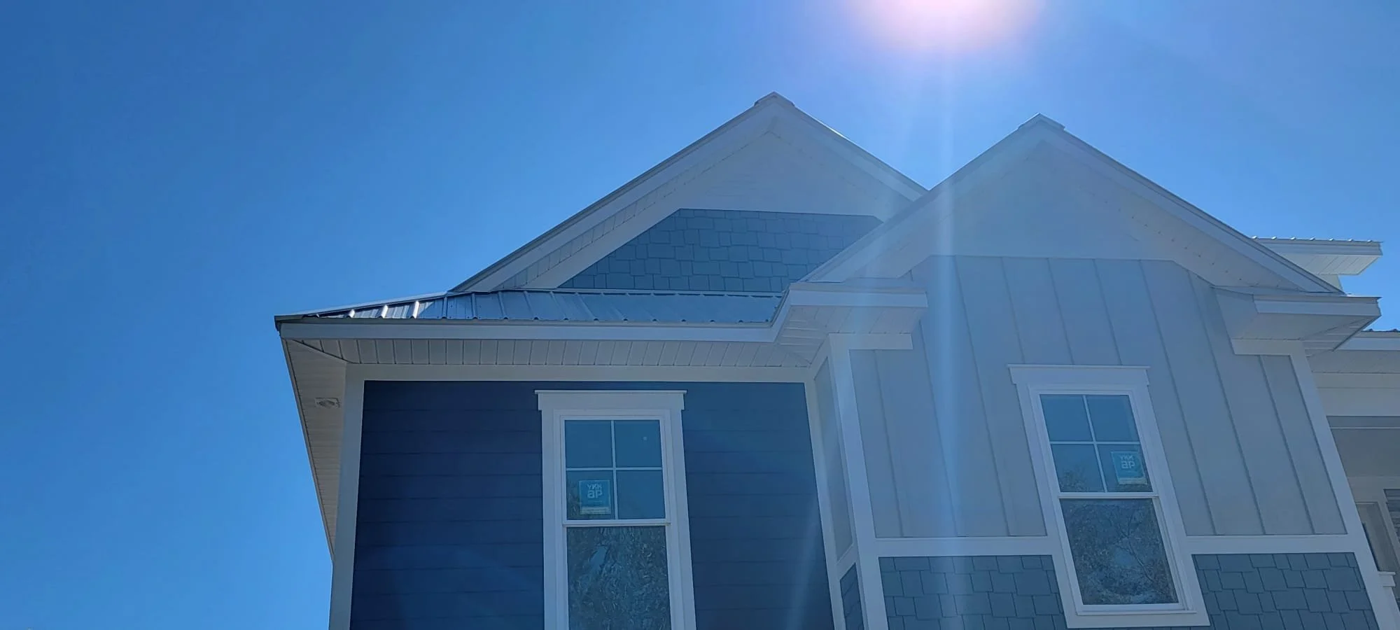 Close-up of a house's corner with a light blue exterior, white trim, and a metal roof, against a clear blue sky with the sun shining from the top right.