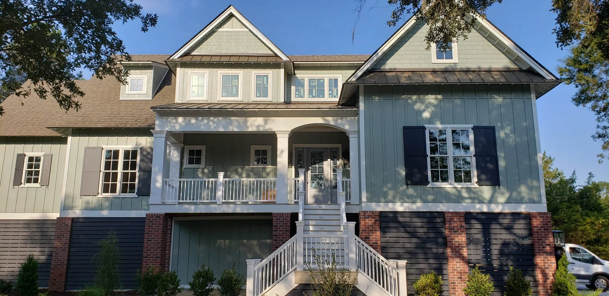 A large, modern house with light green vertical siding, brick accents at the bottom, and black shutters on some windows. The house has a front porch with white railings and stairs leading up to the entrance, surrounded by greenery and trees.