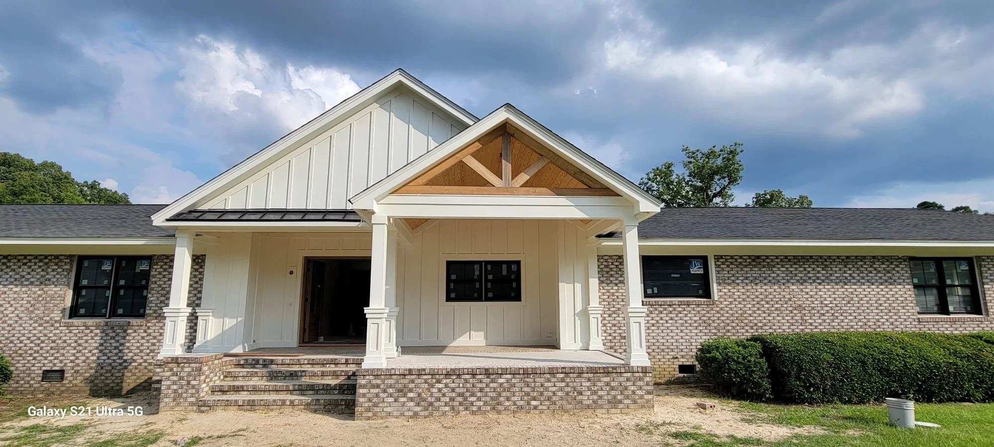 Front view of a new house under construction with a brick lower wall, white vertical siding upper walls, a covered porch, and a gable roof with dark shingles, against a partly cloudy sky.