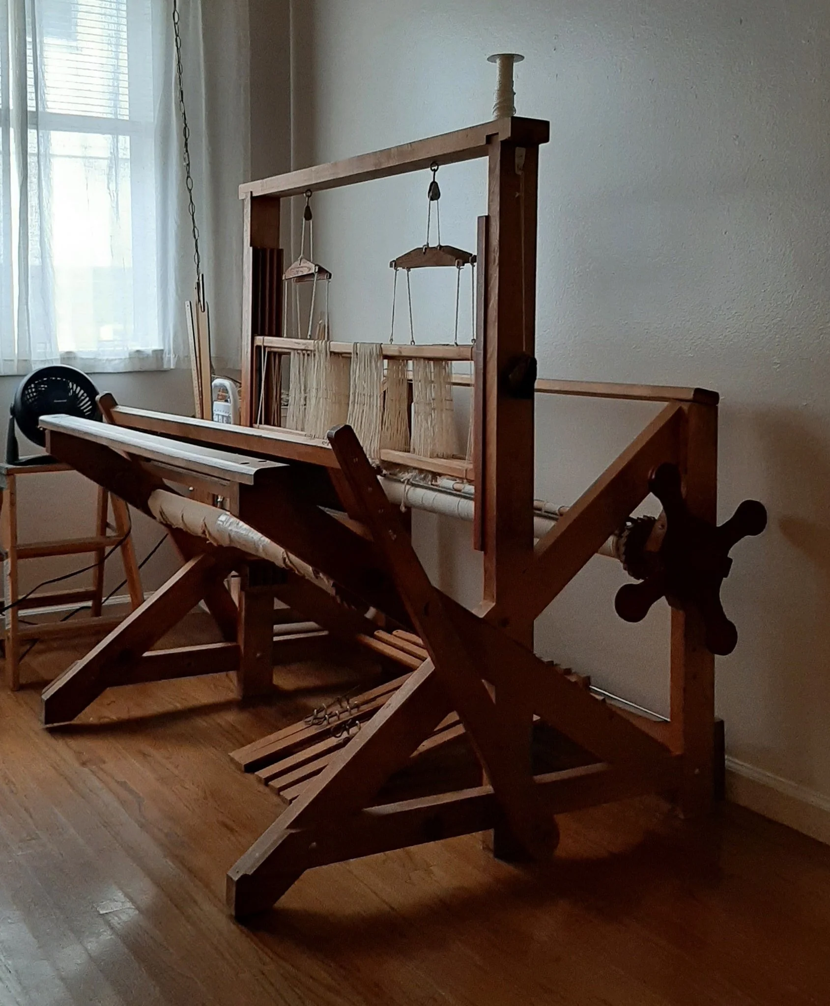 Wooden weaving loom in a room with a wooden floor and a window, creating a warm, traditional craft atmosphere.