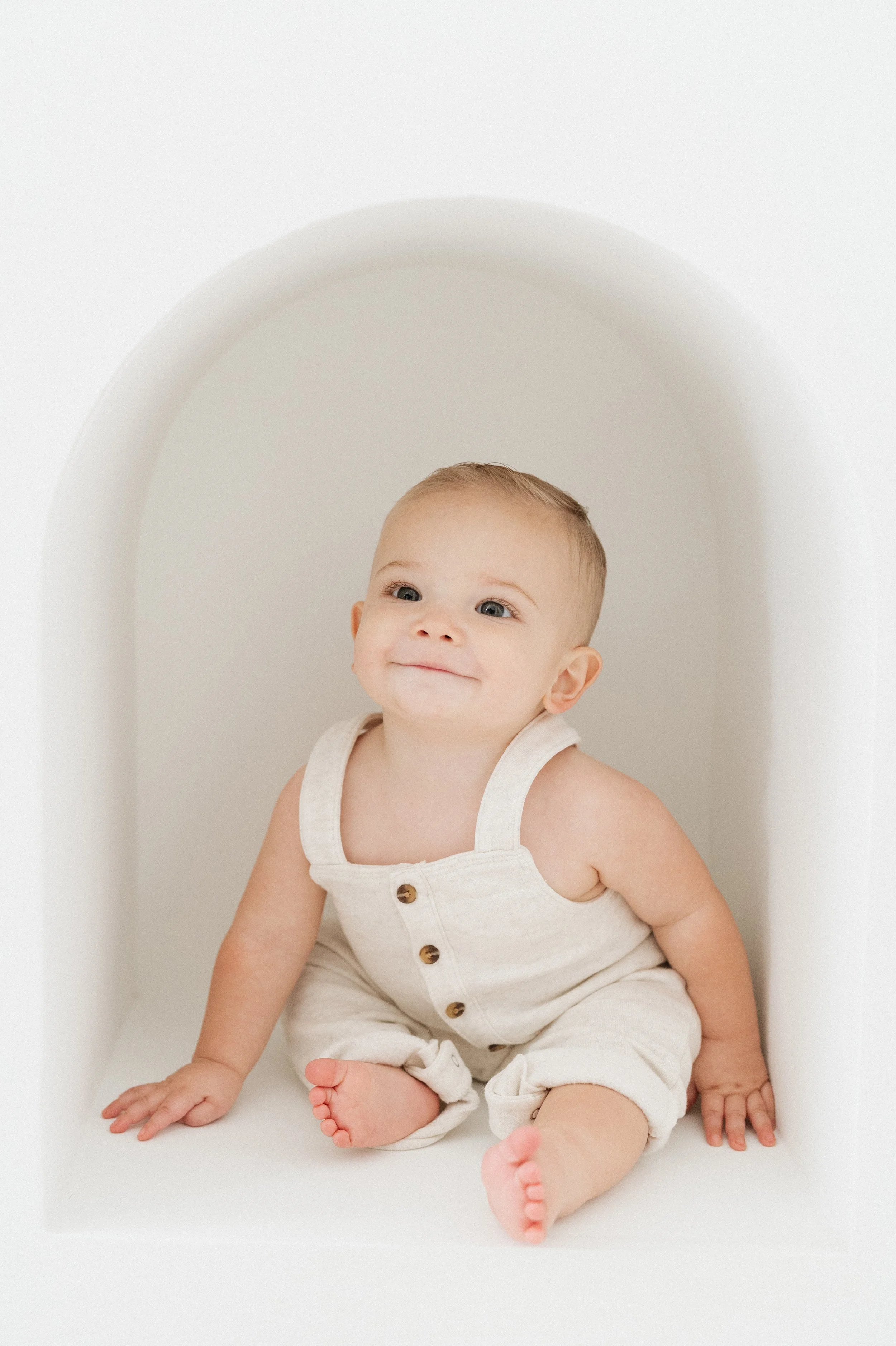 A smiling baby with light skin and blue eyes sitting inside a white arched nook, wearing a sleeveless beige romper with wooden buttons.