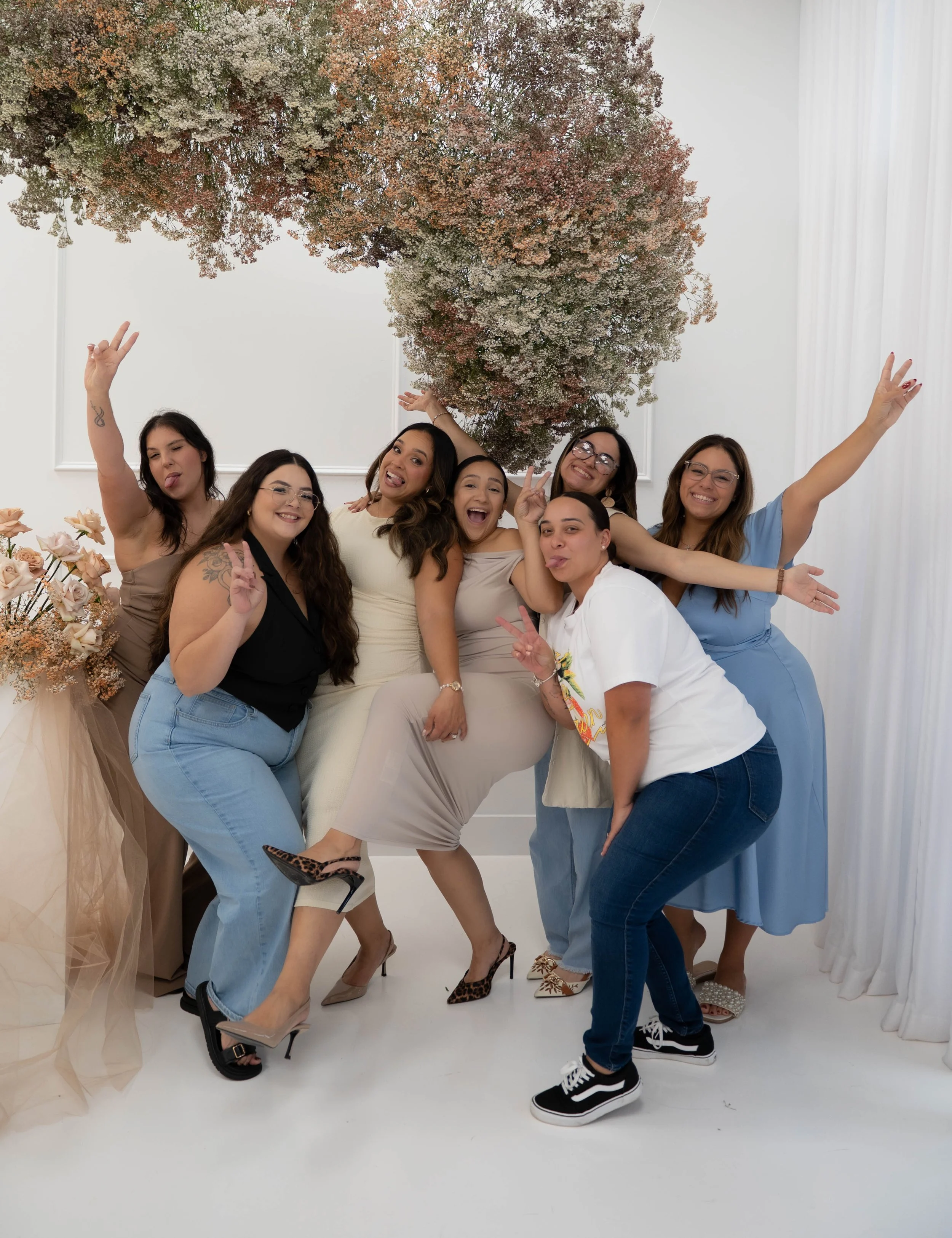 A group of seven women posing and making playful gestures in front of a flower installation with a large, colorful floral arrangement.