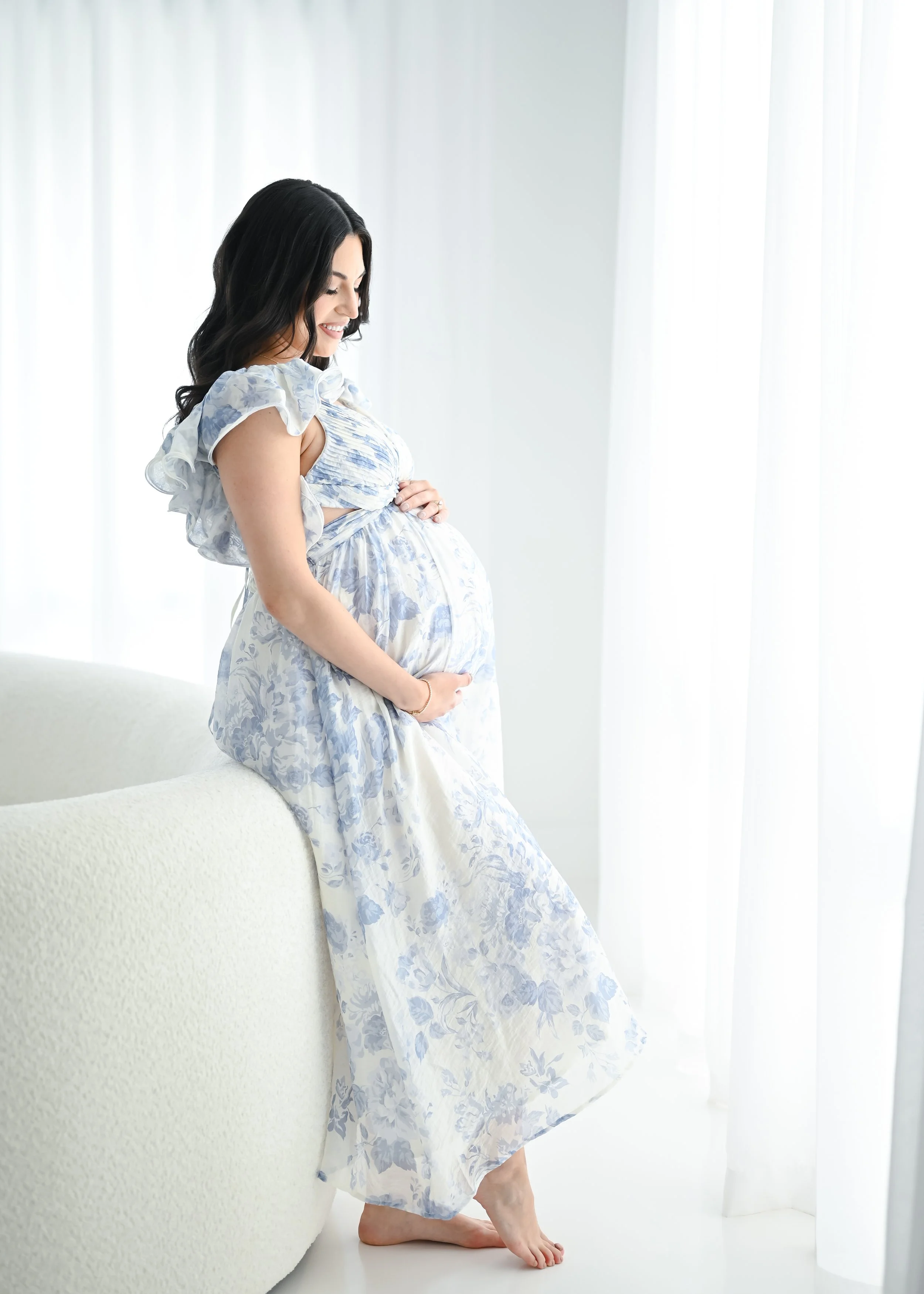 A pregnant woman with dark hair sitting on a white couch, smiling, in a bright room with white curtains, wearing a light blue and white floral dress.