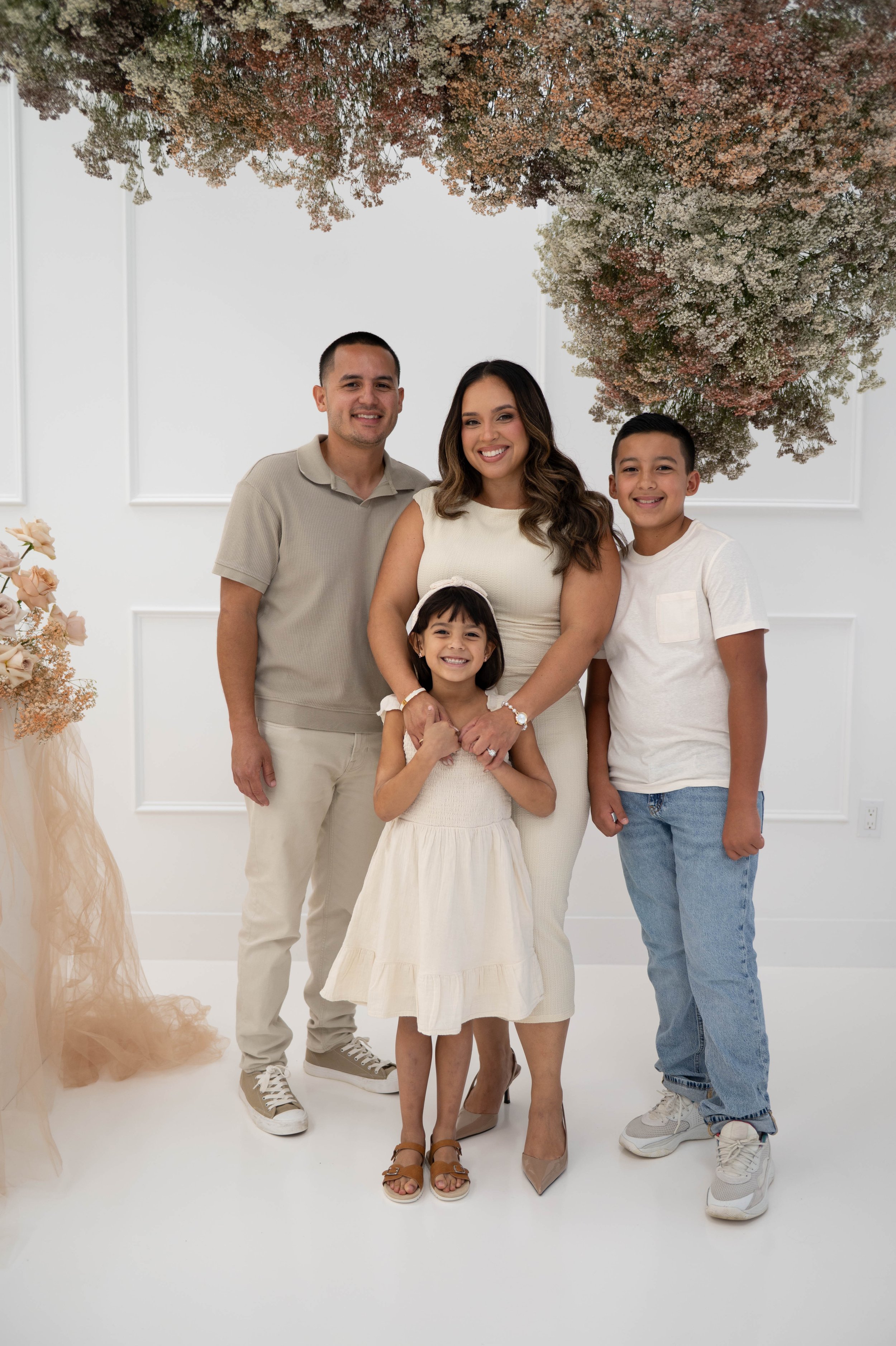 A smiling family of five posing together indoors with white walls and floral decorations, including a large, pink and white floral arrangement hanging from the ceiling.