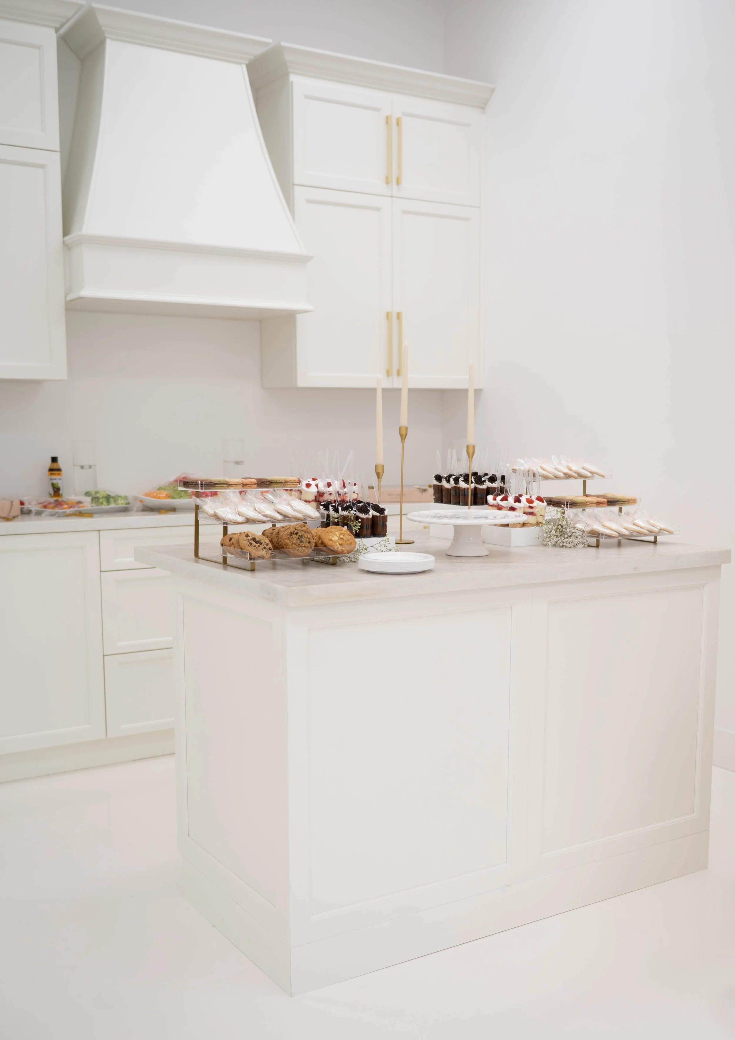 A white kitchen with a dessert table featuring cookies, cupcakes, and cookies on sticks, with candles and decorative accents.