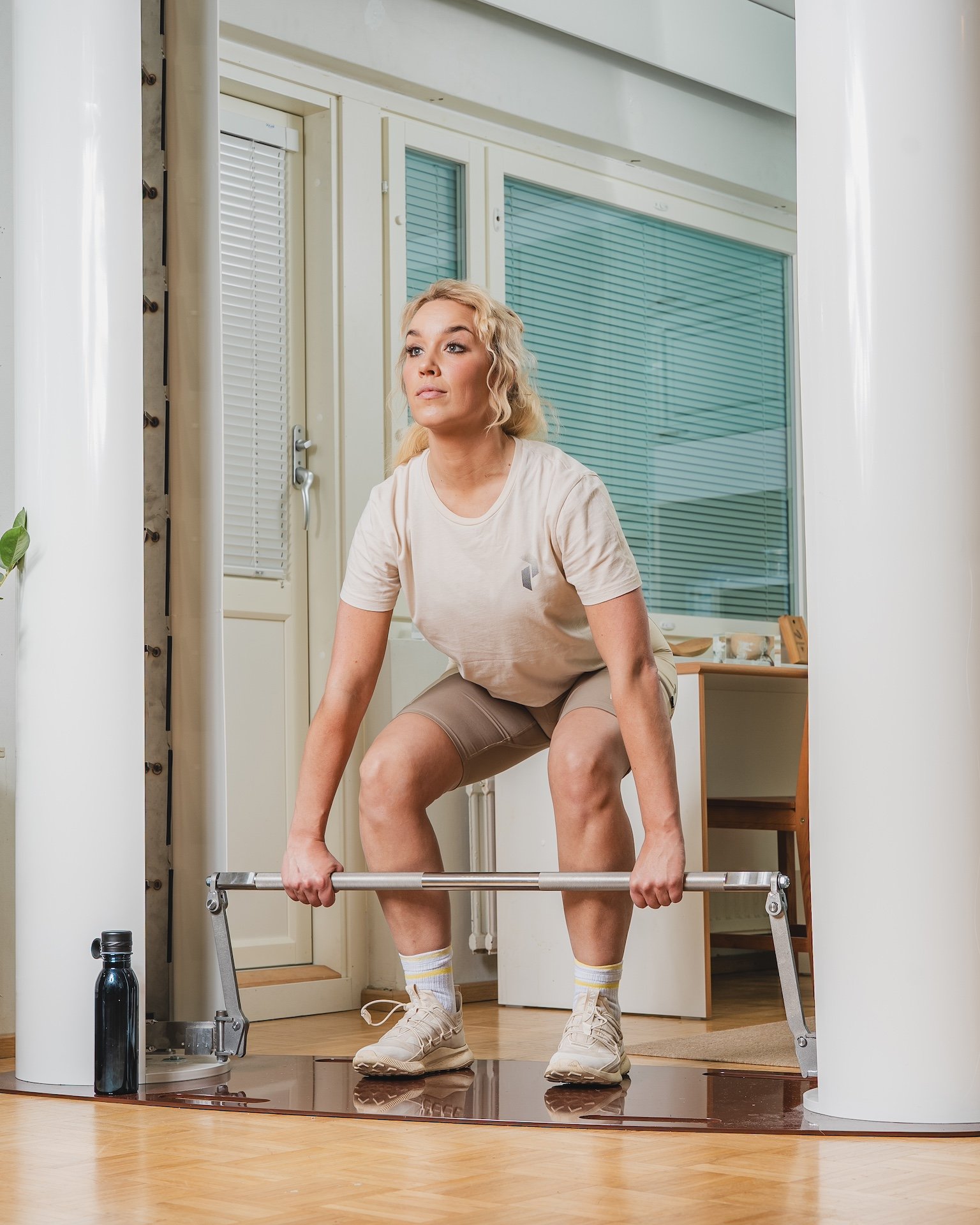 Person performing a deadlift with a barbell in a modern bright room, wearing a beige T-shirt and shorts, with a water bottle nearby.
