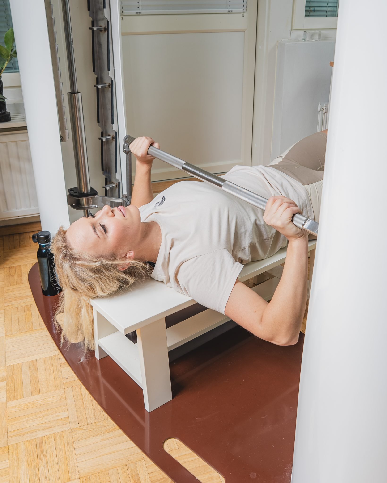 Woman performing a bench press exercise on a white bench with a barbell in a gym setting.