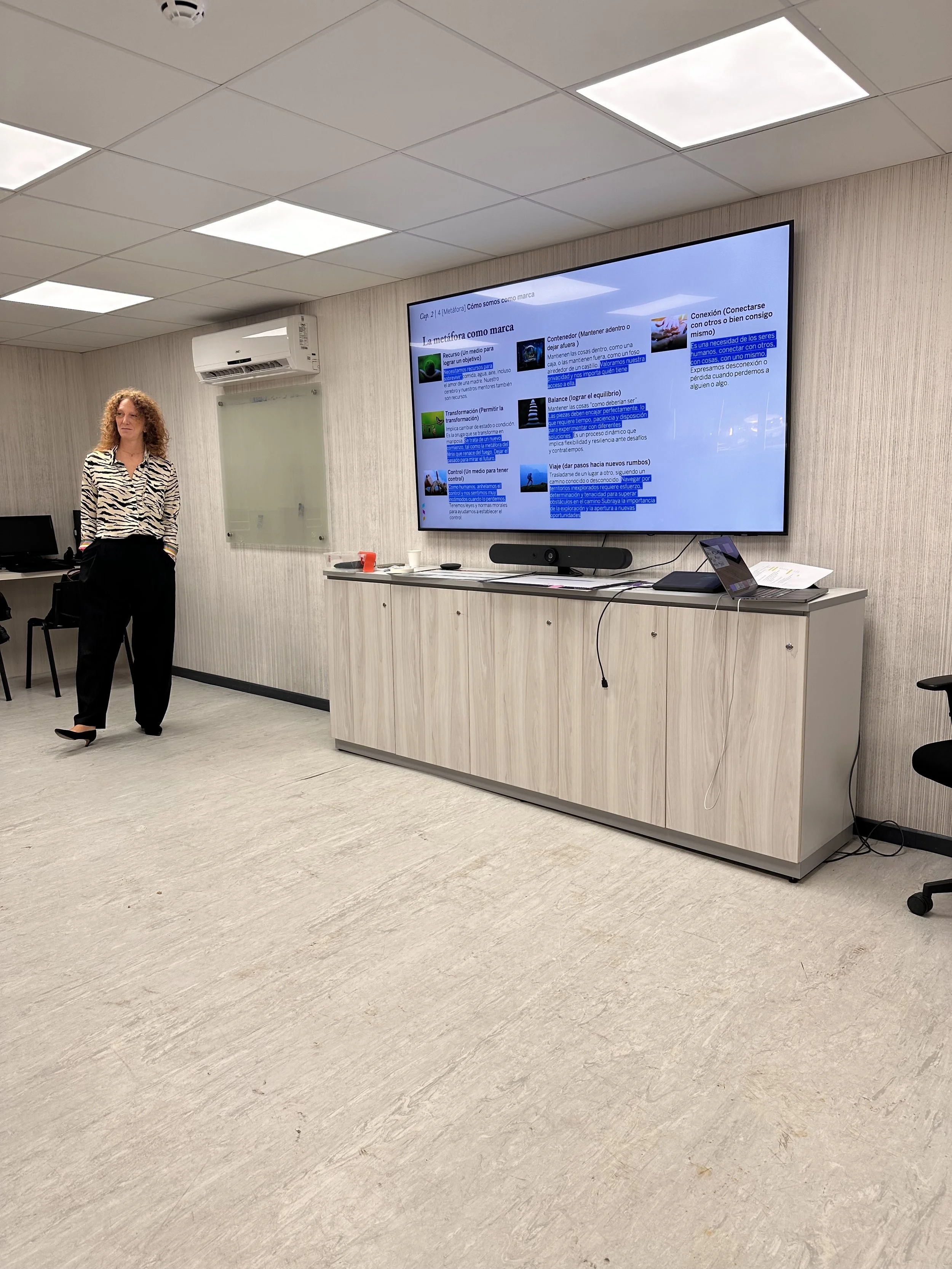 Una mujer de cabello rizado de pie en una sala de conferencias frente a una pantalla grande que muestra una presentación. La sala tiene paredes de color claro, un sistema de aire acondicionado y un mueble con varios equipos electrónicos y documentos.