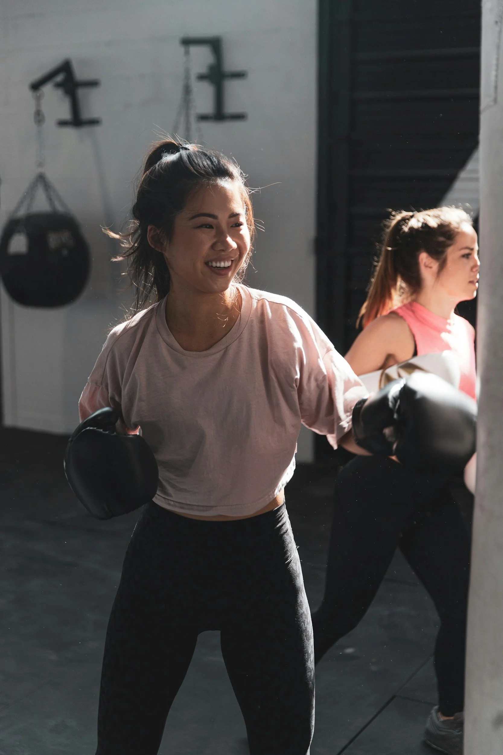 Two women boxing in a gym, wearing casual sports outfits, and black boxing gloves. One is striking a punching bag while smiling.