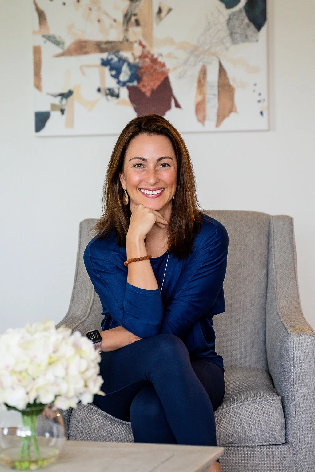 A woman sitting on a gray armchair, smiling, wearing a blue shirt. She has her hand resting under her chin and is wearing a watch and bracelet. A vase with white flowers is on a table in the foreground, and there's abstract art on the wall behind her.