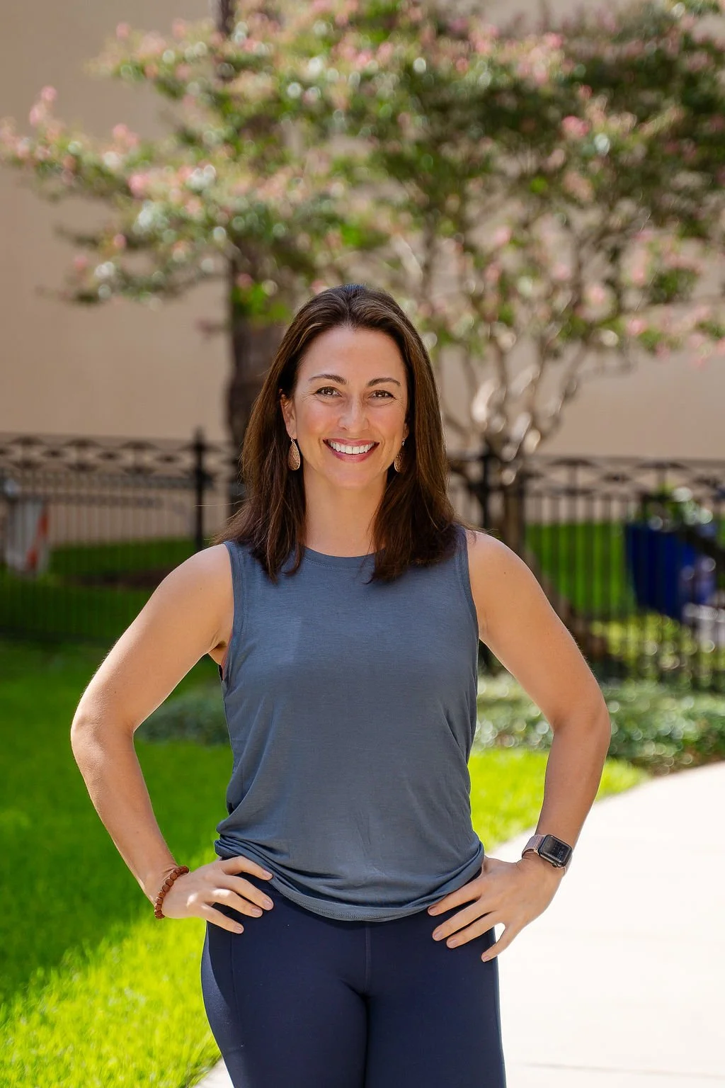 A woman standing outdoors with hands on hips, wearing a sleeveless gray top and dark pants. She is smiling and standing on a pathway with grass and a tree in the background.