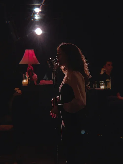A woman singing into a vintage microphone on a dimly lit stage, with a pink lamp and candles in the background.