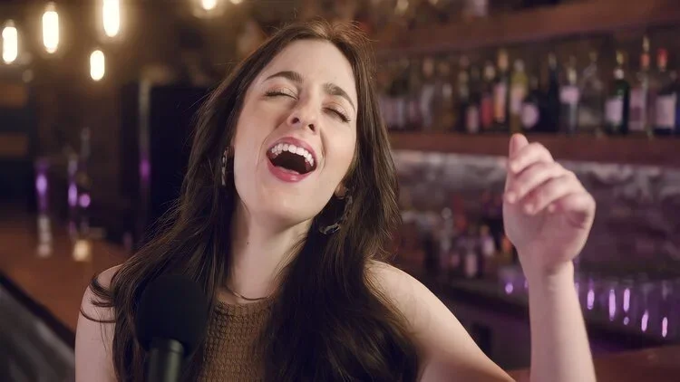 A young woman with long brown hair singing passionately into a microphone in a bar or club setting.