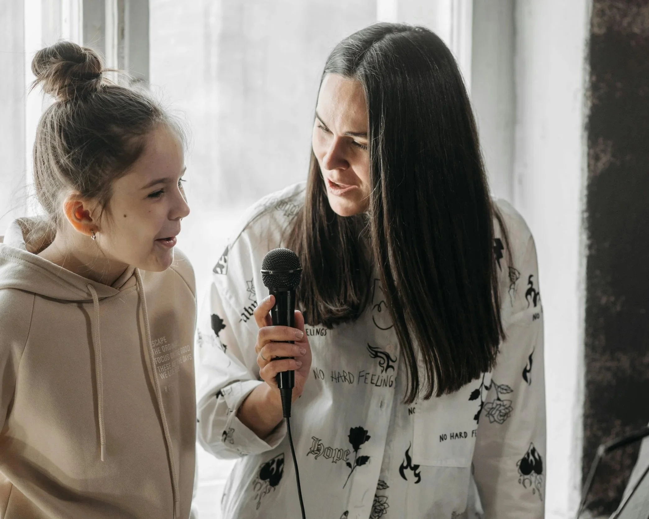 A woman with long dark hair holding a microphone and looking at a young girl with a bun hairstyle. The girl is smiling and talking. They are indoors near a window.
