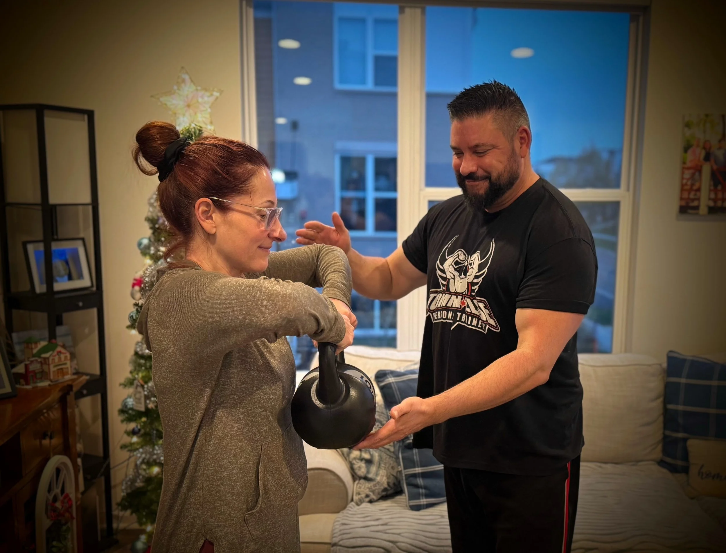 A woman holding a kettlebell with her in-home personal trainer Tommy Else assisting or guiding her during a fitness activity inside a living room with a Christmas tree in the background.