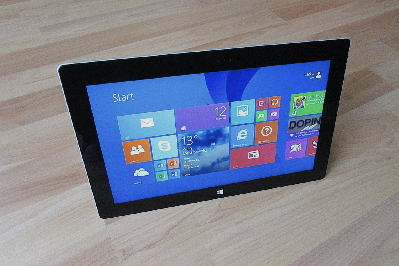 A tablet device displaying the Windows start screen, placed on a light wooden floor.