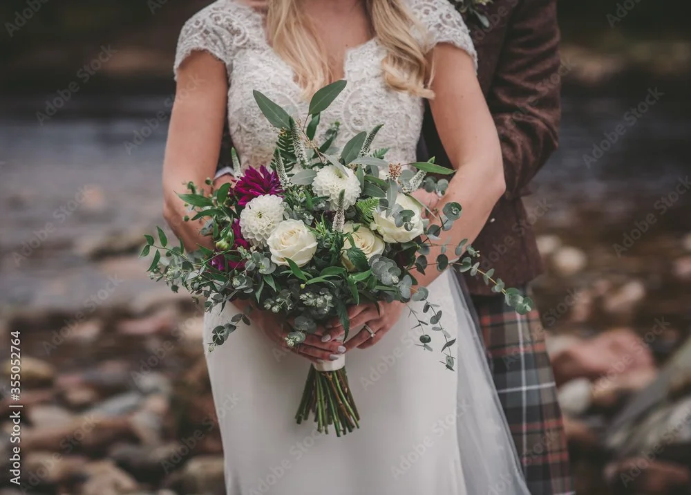 A bride holding a bouquet of white and purple flowers with green foliage, standing outdoors on a rocky surface.