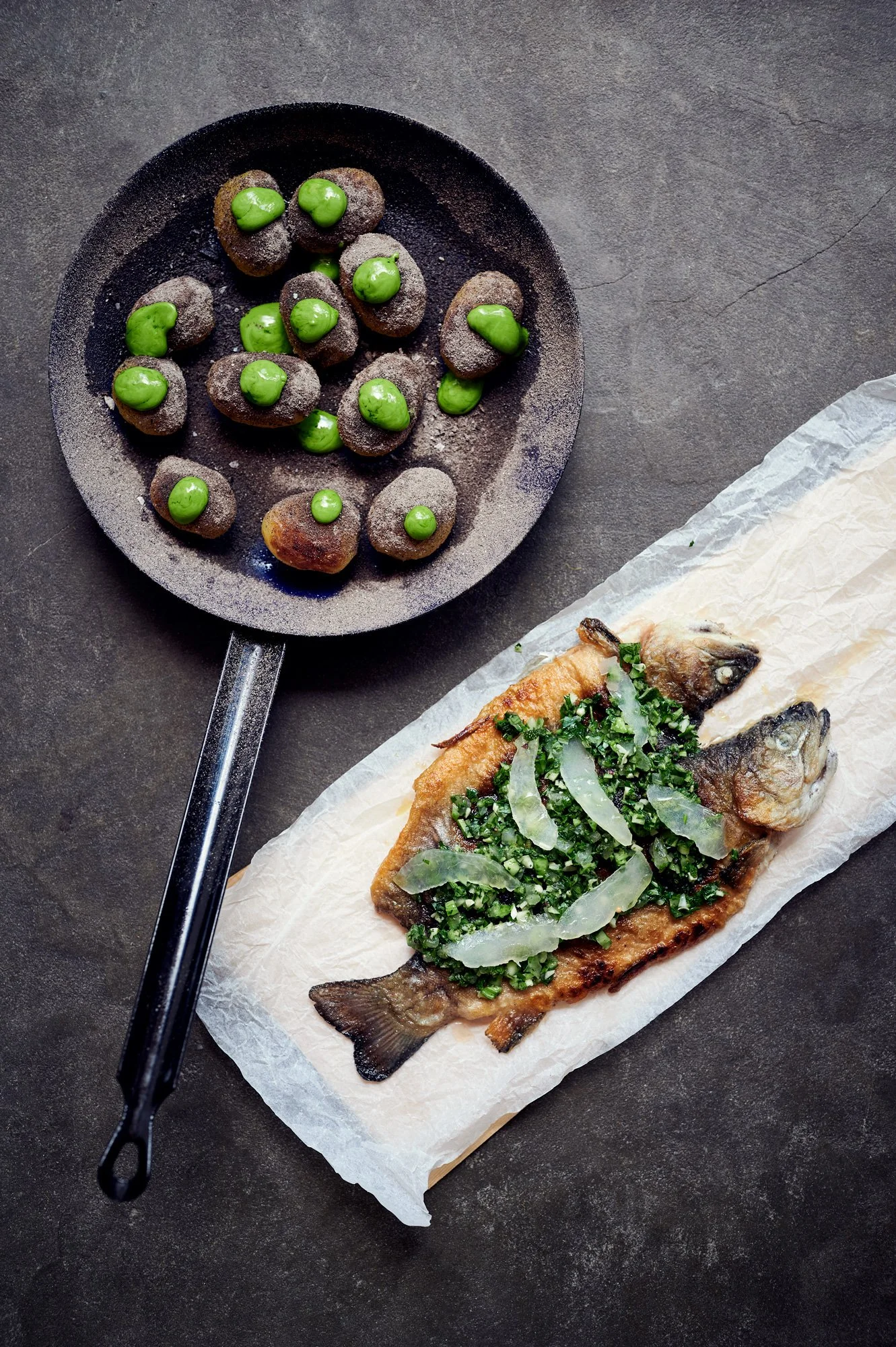 Gegrillter Fisch mit gedünstetem Gemüse auf Backpapier, neben einer Pfanne mit Kartoffelbällchen mit grüner Sauce