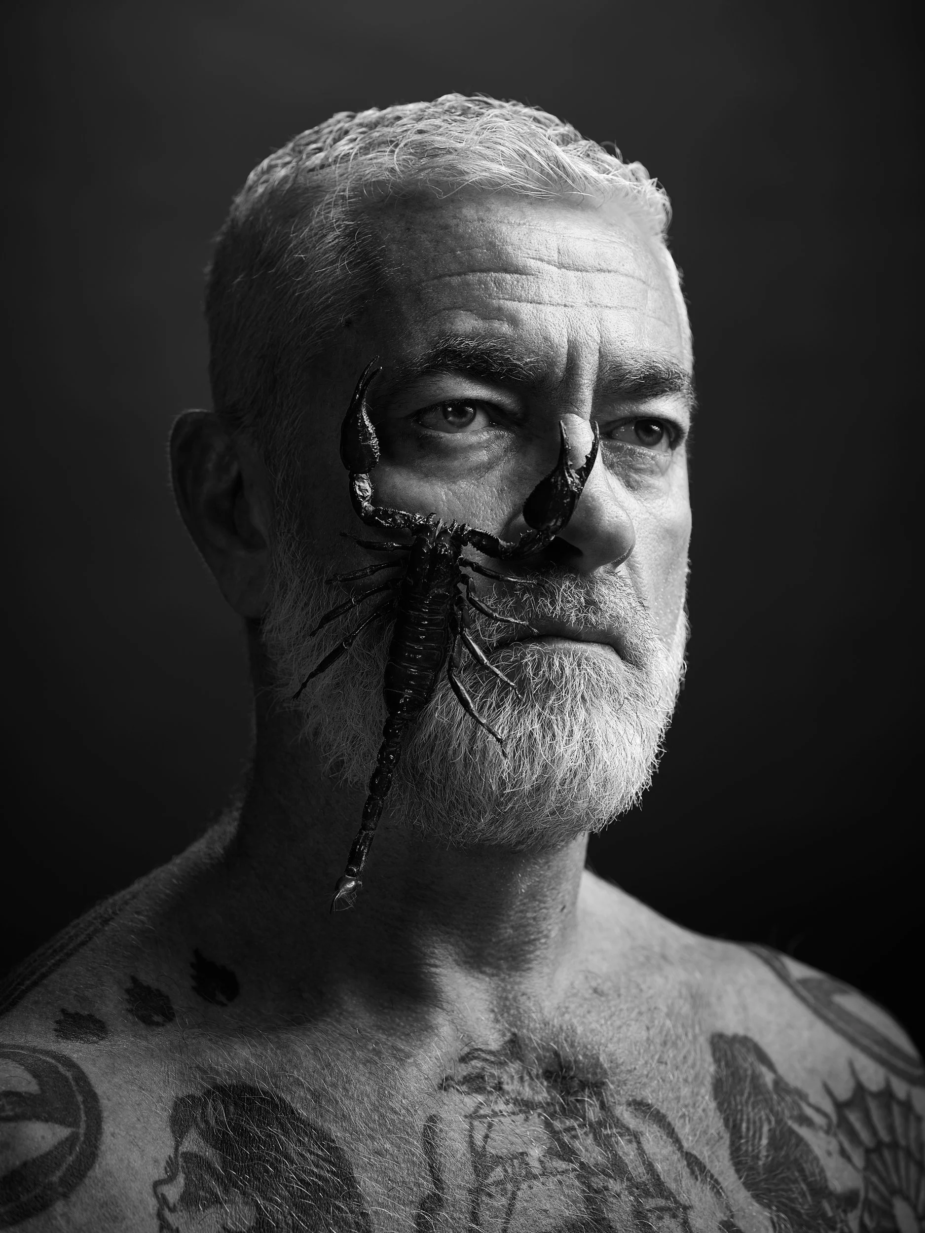 Schwarz-weiß Foto eines Mannes mit Bart und Tattoos, der einen Skorpion auf seinem Gesicht hat.