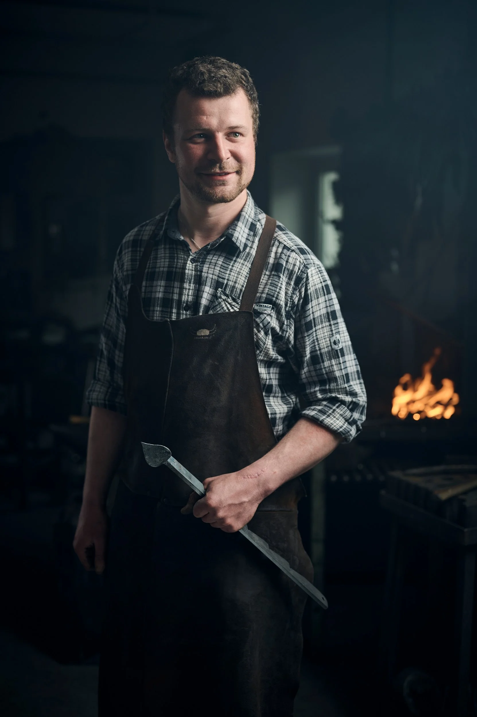 Ein Mann mit kurz lockigem Haar und Bart, trägt eine kariertes Hemd und eine schwarze Schürze, steht in einer dunklen Schmiede mit Feuer im Hintergrund, hält ein Werkzeug in der Hand.