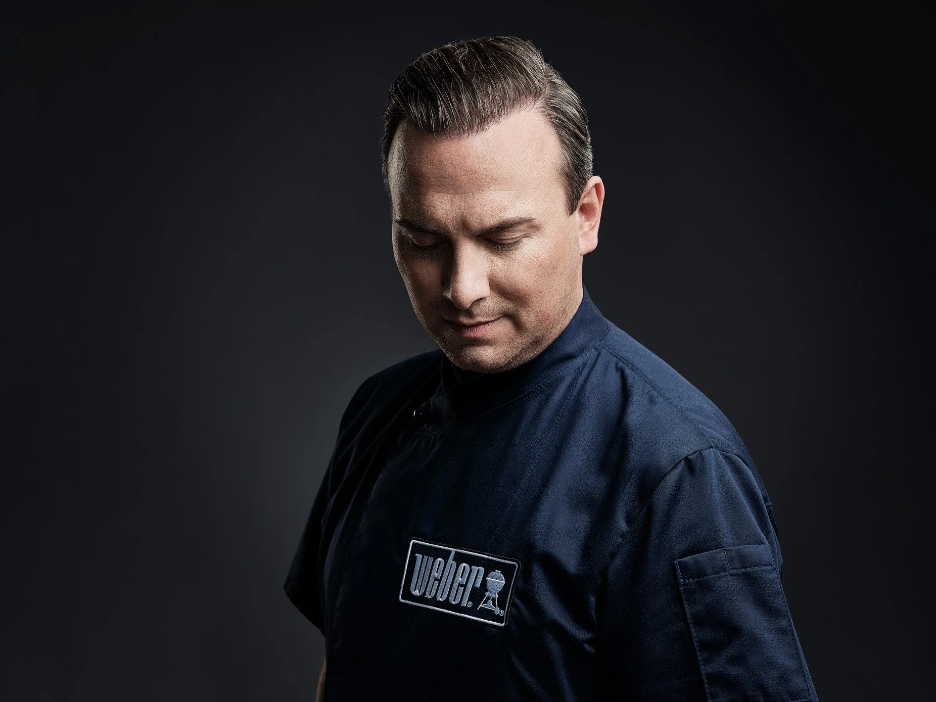 Mann in schwarzem Hemd mit "Weber"-Logo, schaut nach unten, dunkler Hintergrund.