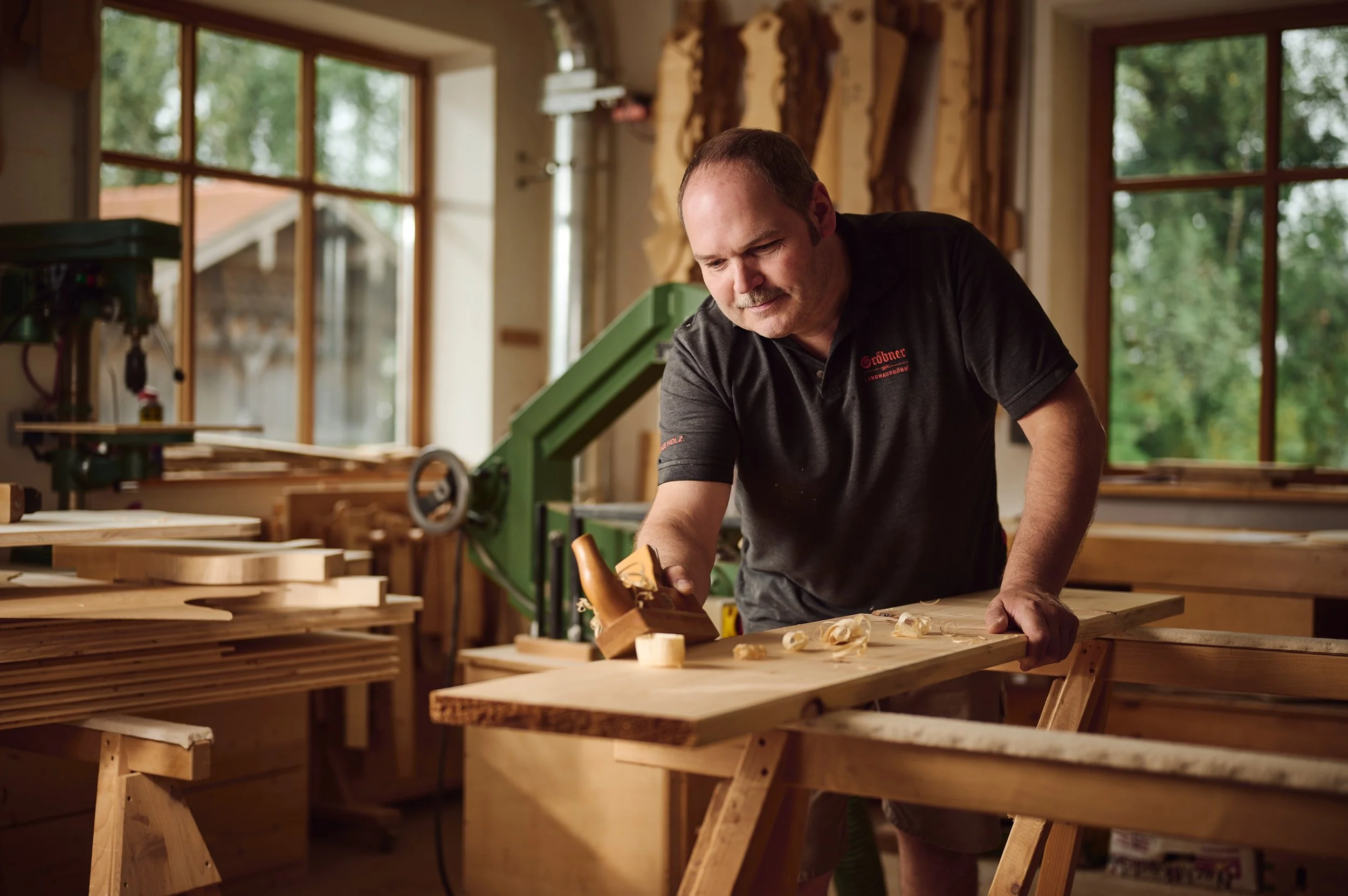 Mann in Werkstatt arbeitet mit Holz an einem Arbeitstisch, umgeben von Holzwerken und Maschinen, große Fenster im Hintergrund.