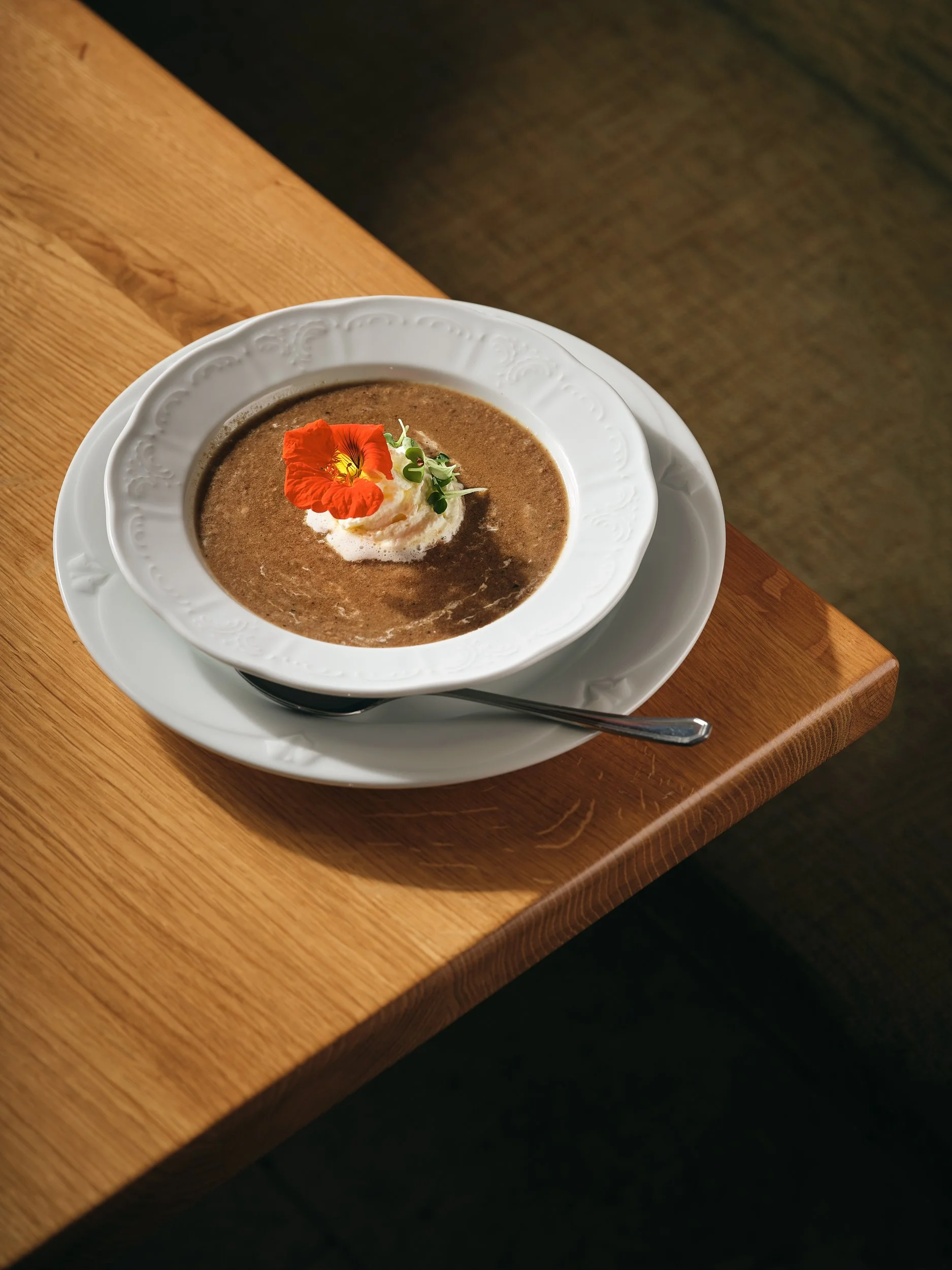 Teller mit Suppe, garnierte mit einer Creme, einer roten Blume und grünen Kräutern, auf einem weißen Teller auf einem Holztisch.