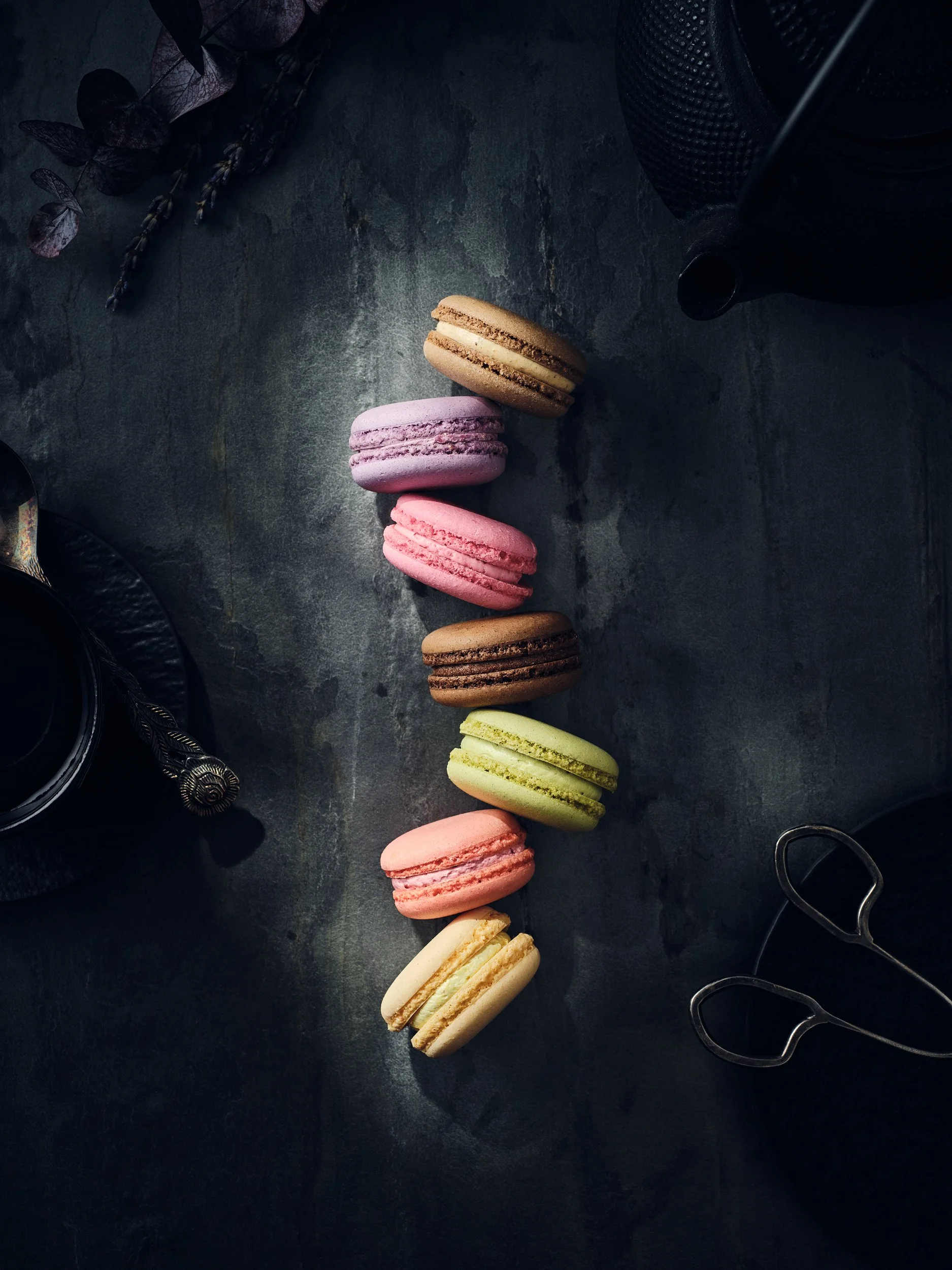 Bunte Macarons auf dunkler Oberfläche, Teekanne und Teetasse im Hintergrund.