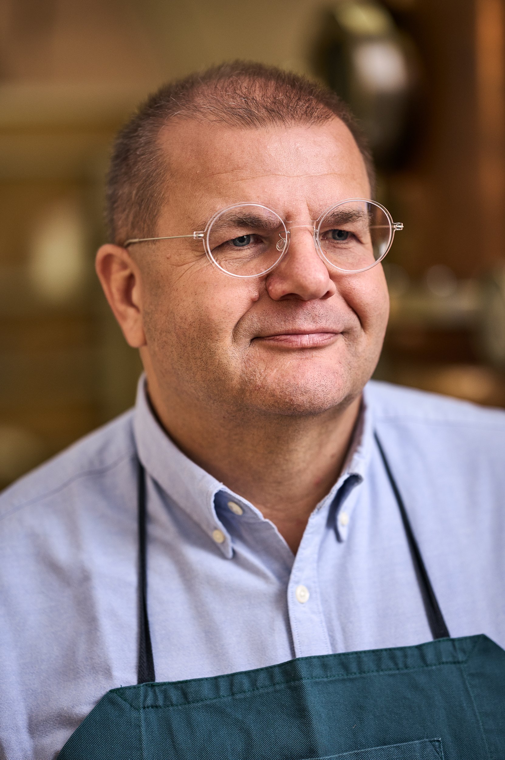 Ein Mann trägt eine Brille mit runden Gläsern und trägt eine graue Hemd. Er trägt eine grüne Schürze und ein Stethoskop um den Hals, sieht freundlich aus.