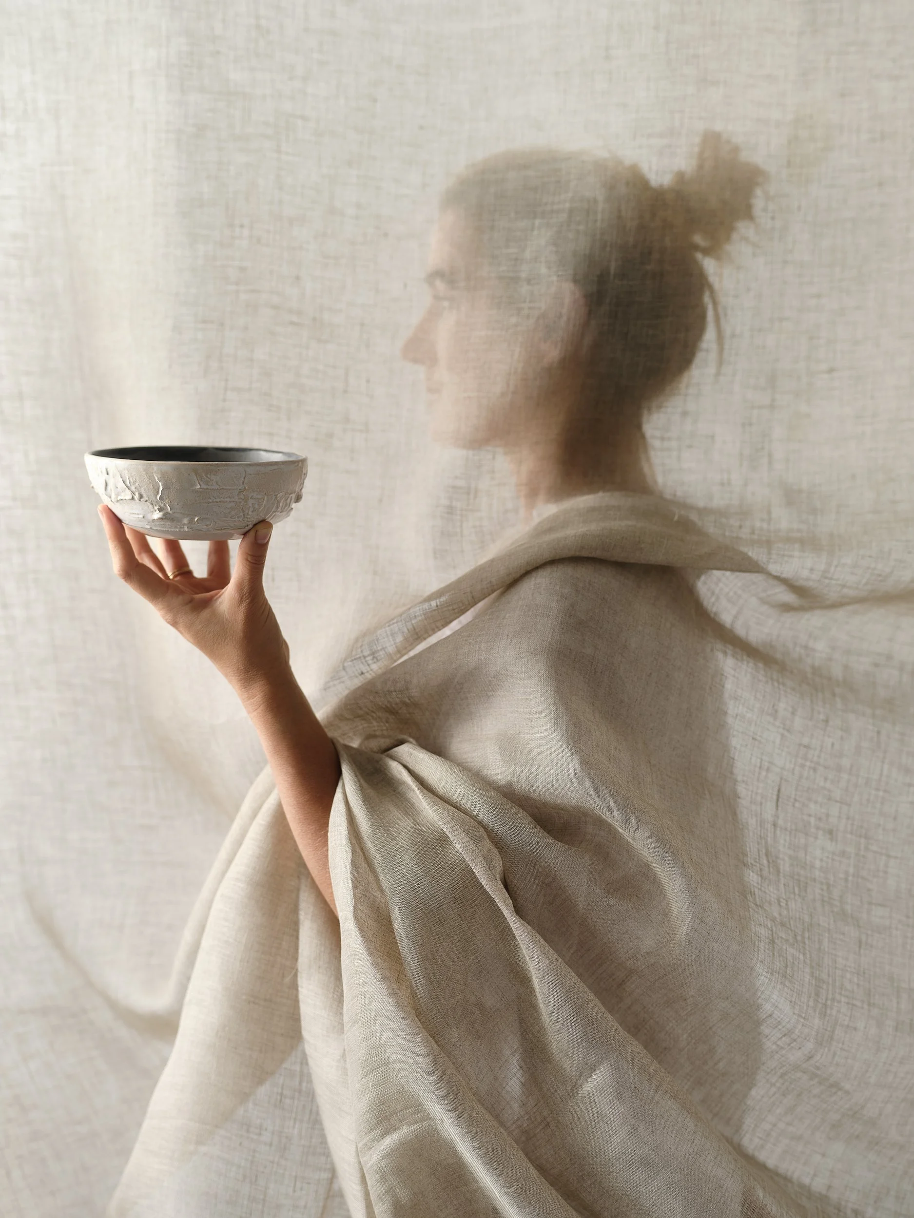 Eine Frau, die hinter einem halbtransparenten beige Stoff steht, hält eine weiße Schüssel. Das Bild ist unscharf und künstlerisch gestaltet.