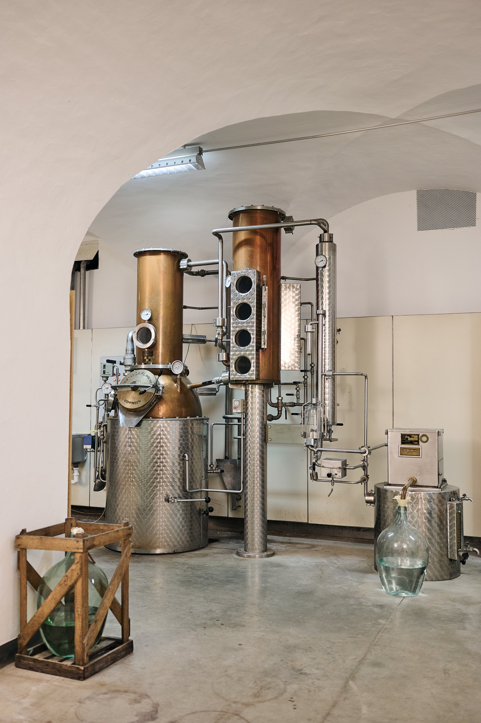 Industrielle Destillationsanlage mit Kupfer- und Edelstahlrohren in einem Laborraum, umgeben von Glasbehältern und einem Holzkasten mit Glasflaschen.
