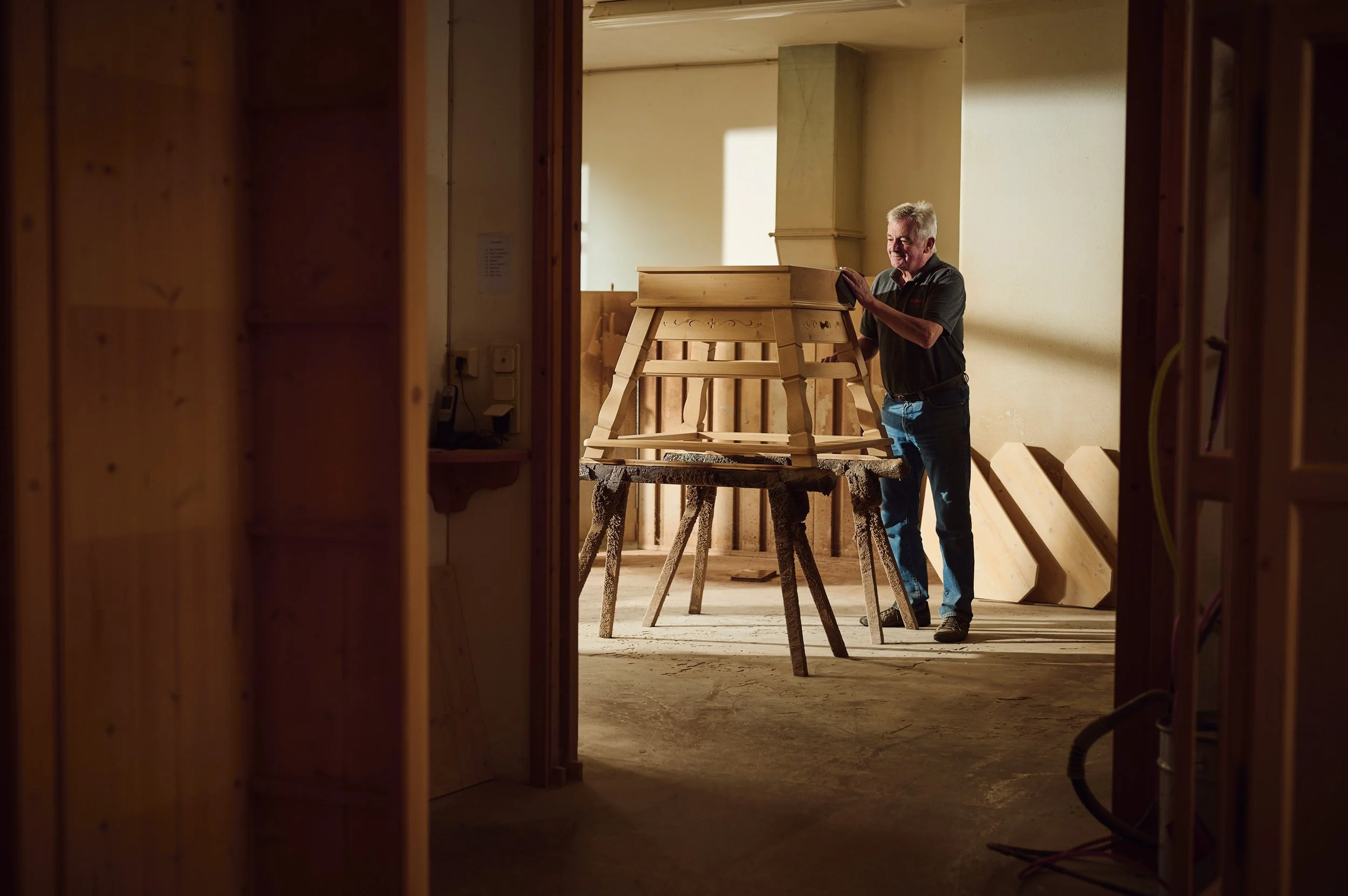 Ein älterer Mann arbeitet in einem Holzhaus, der Blick wird durch eine Türöffnung auf einen Raum mit Holzarbeiten und -strukturen gerichtet.