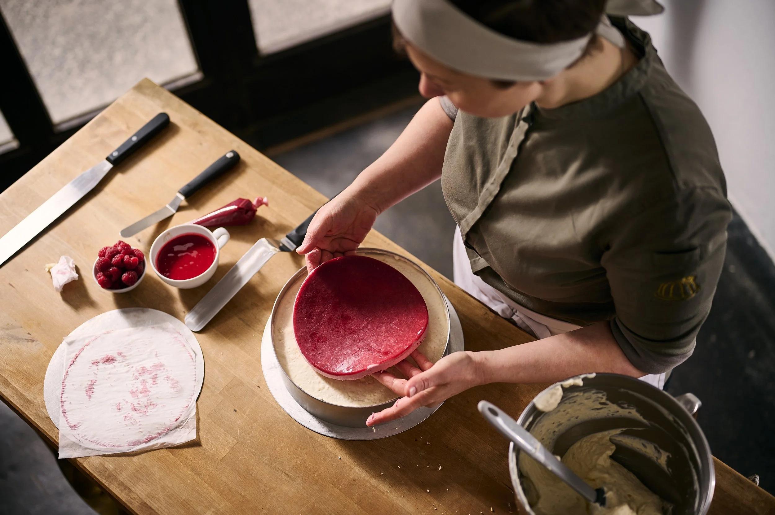 Eine Person bereitet eine rote Schicht für eine Torte vor, während auf einem Tisch ein Schneide Messer, frische Himbeeren, eine Schüssel mit roter Fruchtfüllung, und andere Backaccessoires liegen.