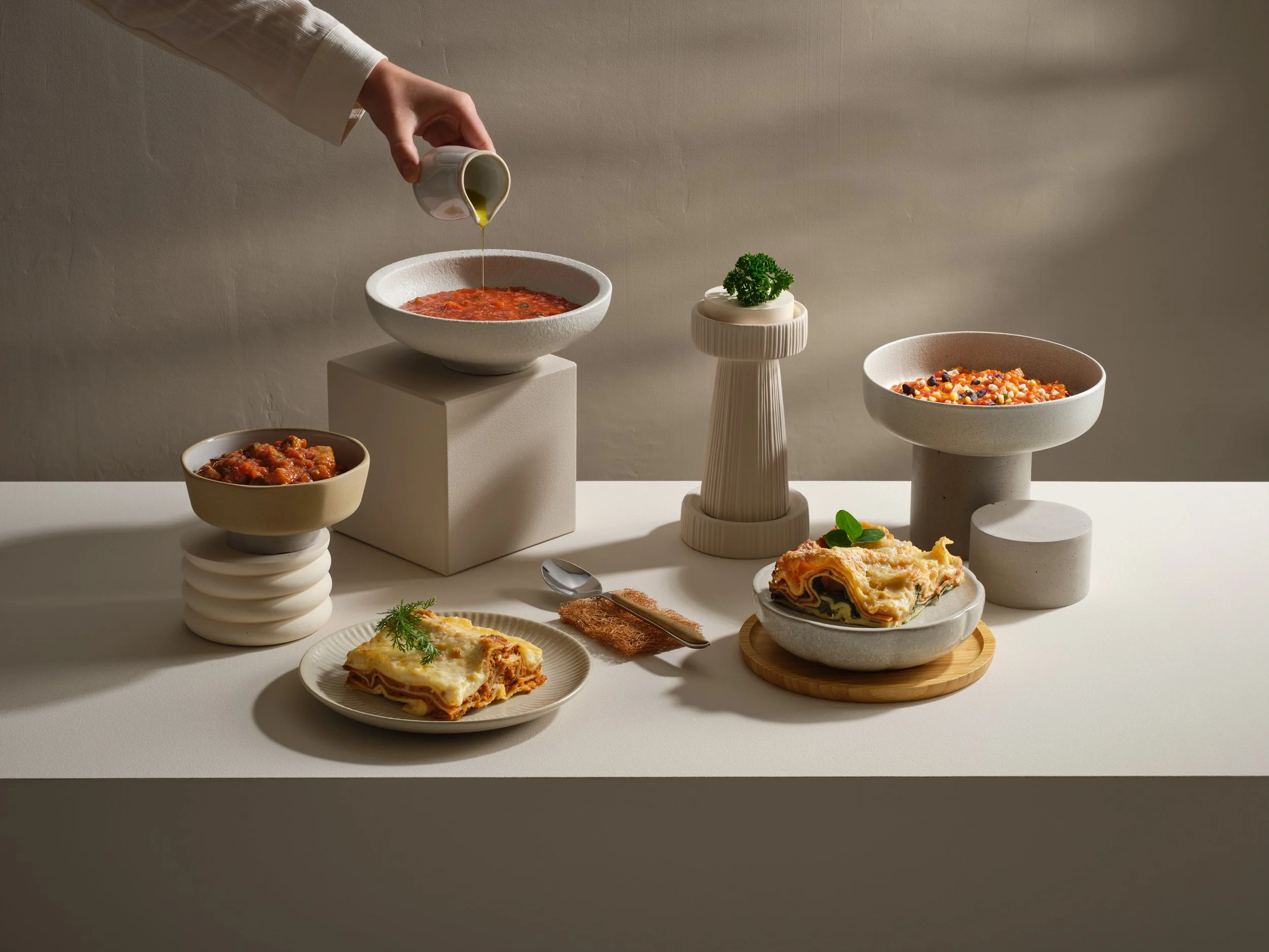 Verschiedene Gerichte und  Küchenutensilien auf einem weißen Tisch, darunter eine Tomatensuppe, Lasagne, ein Salat, und ein Stück Kuchen, mit minimalistischen keramischen Dekorationen im Hintergrund.