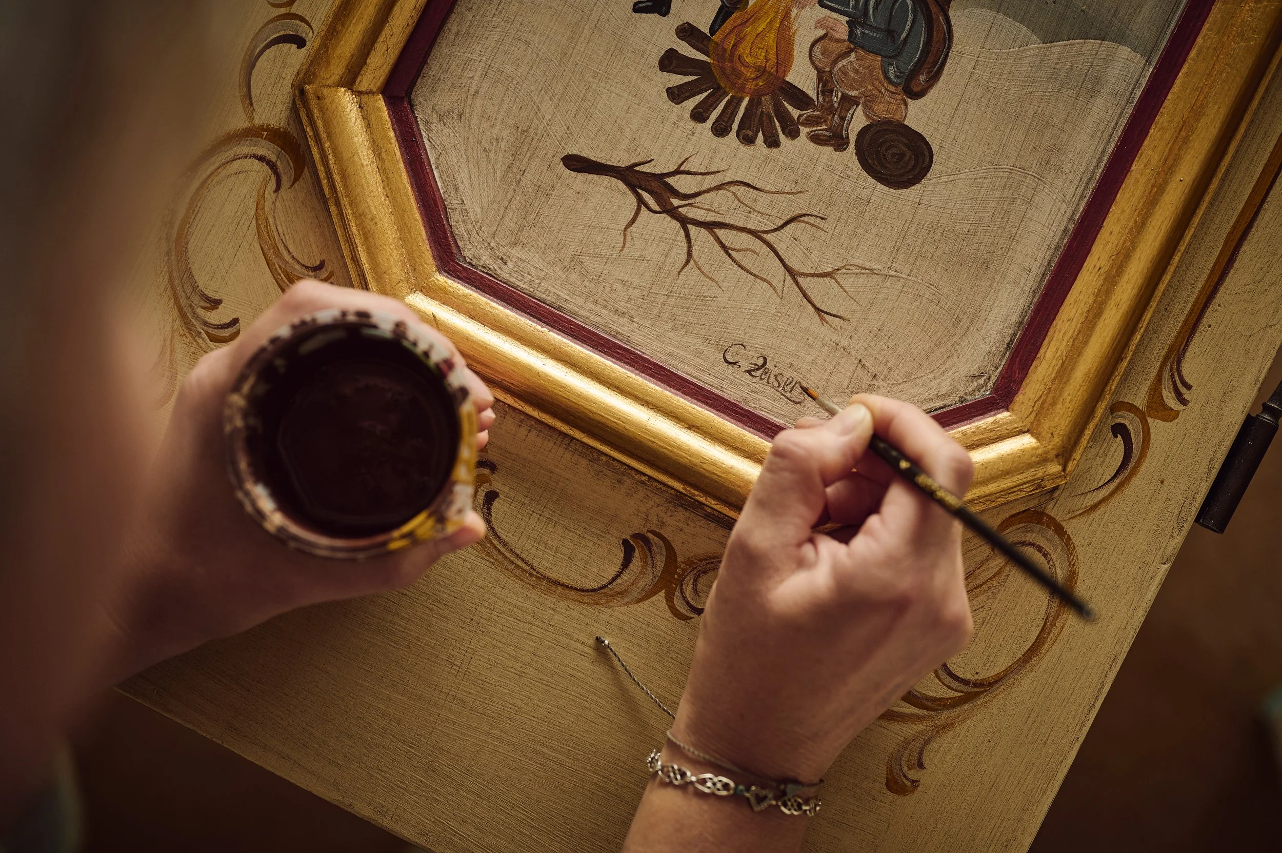 Ein Künstler trägt Goldfarbe auf eine schwere, quadratische Holztafel mit einem Pinsel auf, die mit einem kunstvollen Rahmen verziert ist. Die Oberfläche der Tafel zeigt eine Grafik, die eine abstrakte Darstellung von Natur und Gegenständen enthält, darunter ein künstlerisch gestalteter Baum, eine spiralartige Form und andere organische Formen. Das Bild ist bei einer Lichtquelle aufgenommen und zeigt die Hand des Künstlers, der eine kleine Dose mit dunkler Farbe hält.