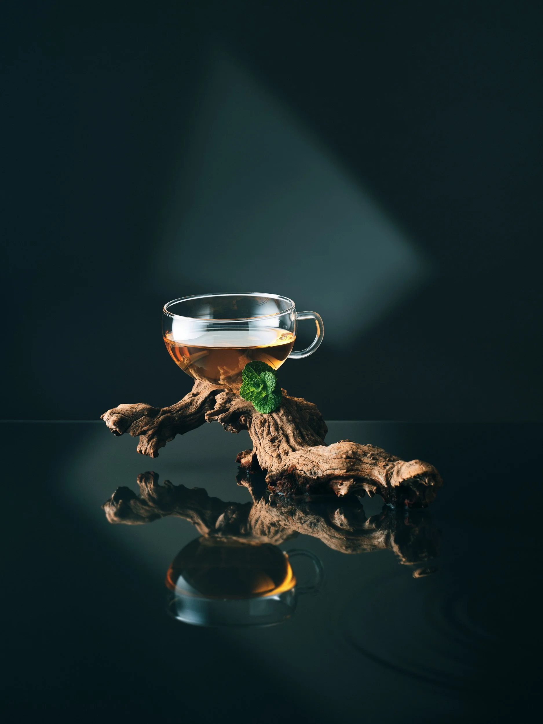 Ein klarer Teetassen mit warmem Tee auf einem Holzstück, mit grünen Minzblättern, auf einem schwarzen reflektierenden Tisch, dunkler Hintergrund.
