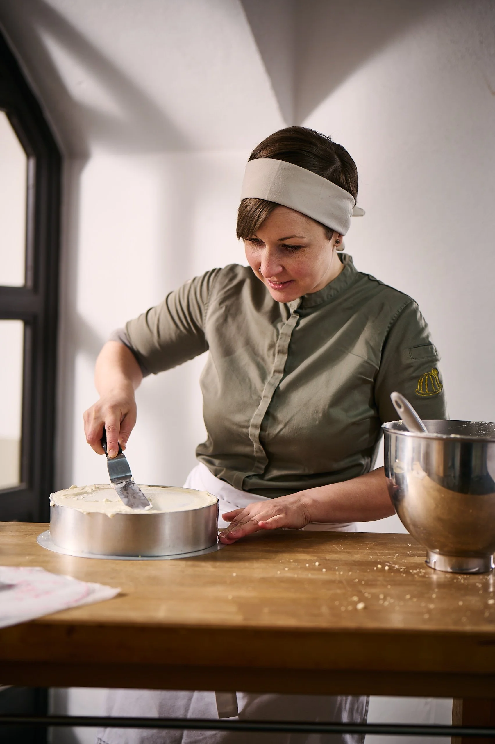 Eine Frau backt einen Kuchen in einer Küche, trägt eine grüne Arbeitsjacke und ein Kopfband, steht hinter einem Holztisch und schneidet den Kuchen.