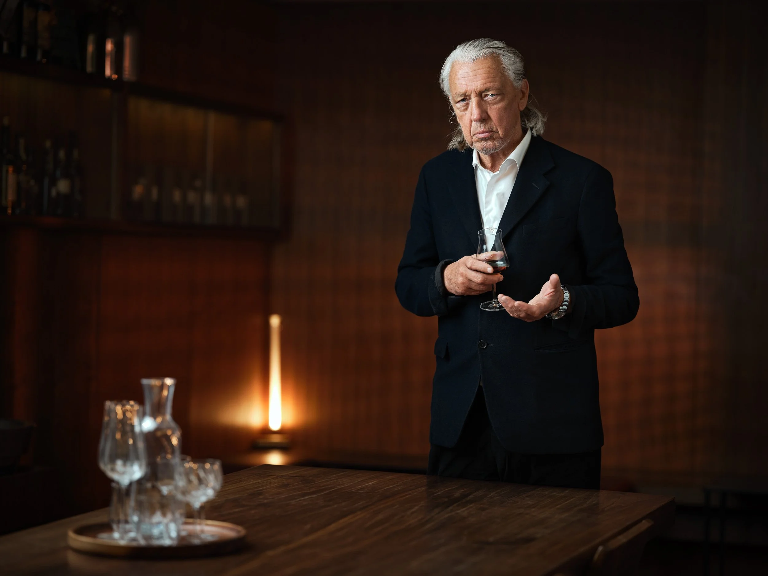 Älterer Mann in Anzug hält ein Glas Schnaps in einem elegant beleuchteten Raum. Im Vordergrund ein Tisch mit leeren Gläsern und einer Karaffe.