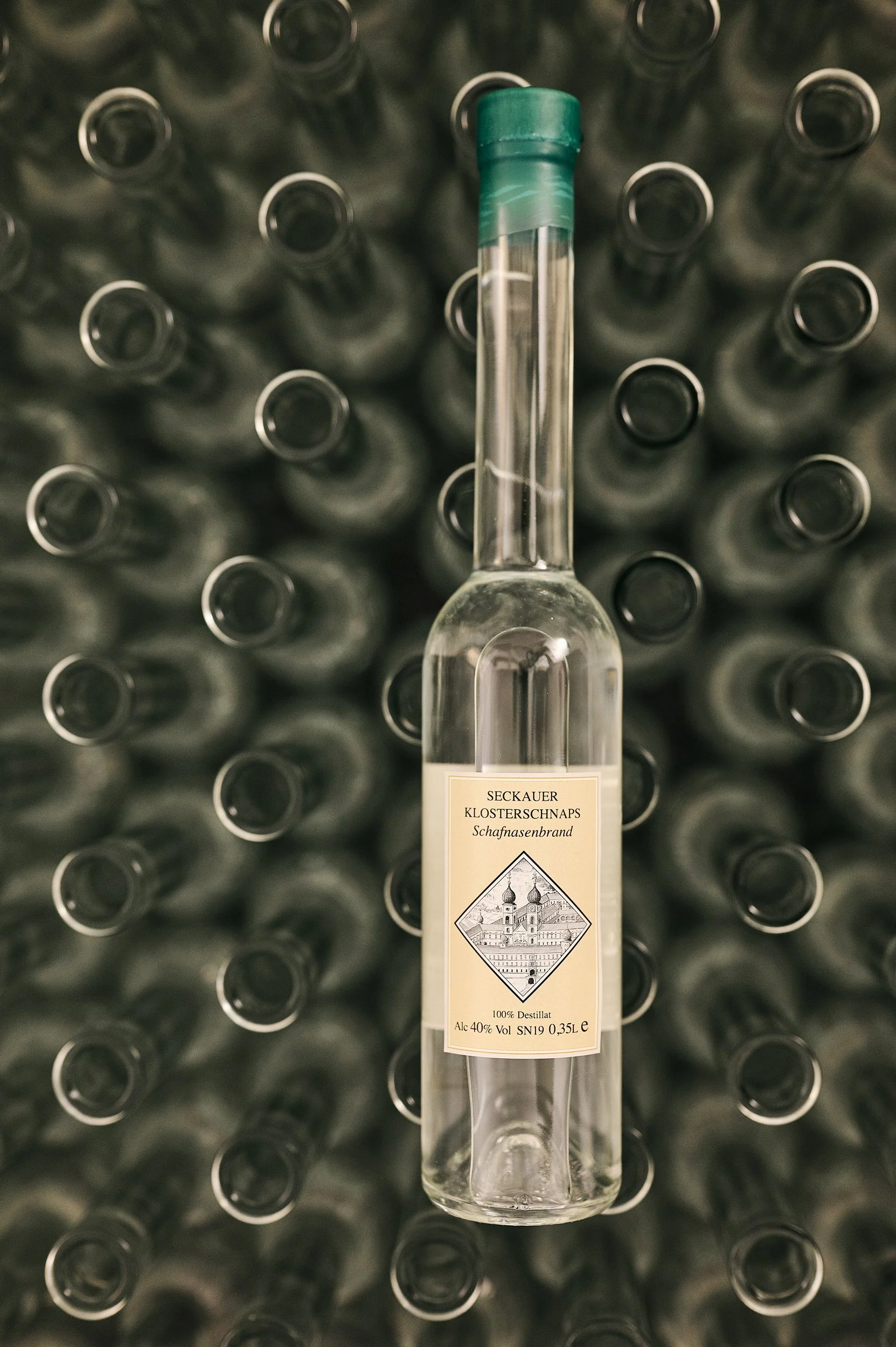 In der Mitte des Bildes befindet sich eine Flasche mit einem Etikett, das auf einen Brand namens 'Klosterschnaps' hinweist. Die Flasche steht vor einem Hintergrund aus vielen kappenlosen Flaschen, die vermutlich leer sind.