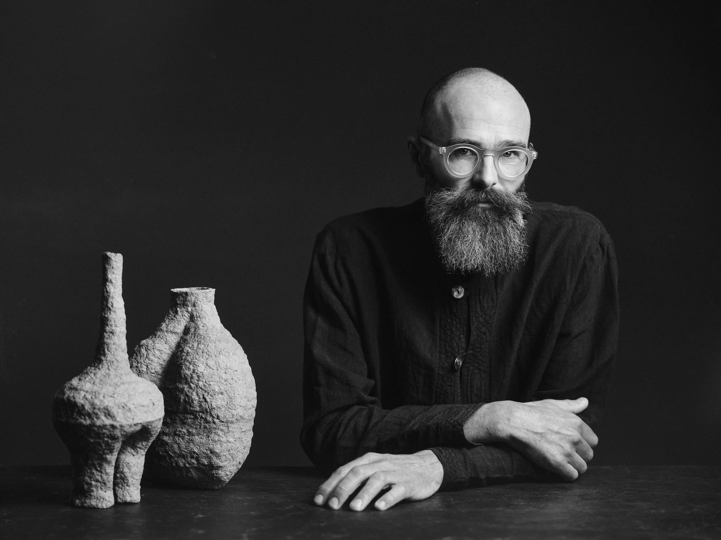 Schwarz-weiß-Fotografie eines Mannes mit Bart, Brille und eleganter Kleidung, der auf einem Tisch sitzt, neben ihm stehen mehrere rundliche, kleine Skulpturen aus Papier.