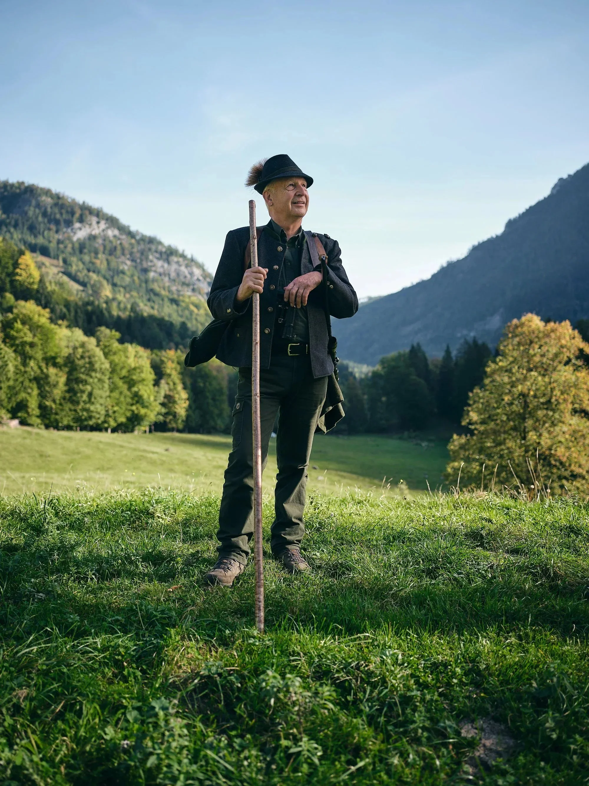 Ein älterer Mann steht auf einer grünen Wiese vor einer bergigen Landschaft, trägt einen schwarzen Hut mit Bommel, eine schwarze Jacke, Hosen und Wanderstiefel, hält einen Wanderstock in der Hand und schaut in die Ferne.