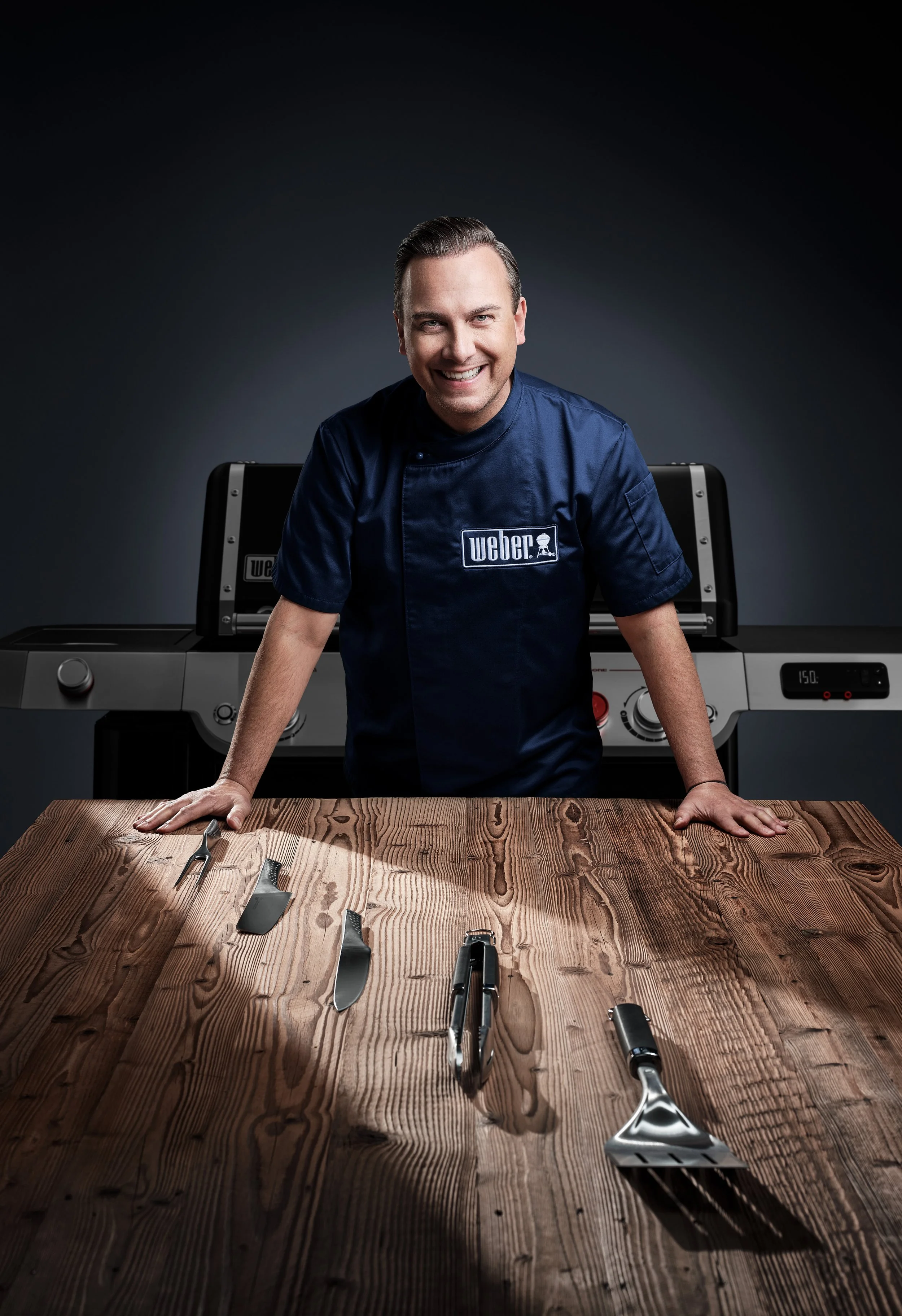 Mann in dunkelblauer Kochuniform steht lächelnd hinter einem Holztisch mit Grillbesteck, im Hintergrund ein Grill.