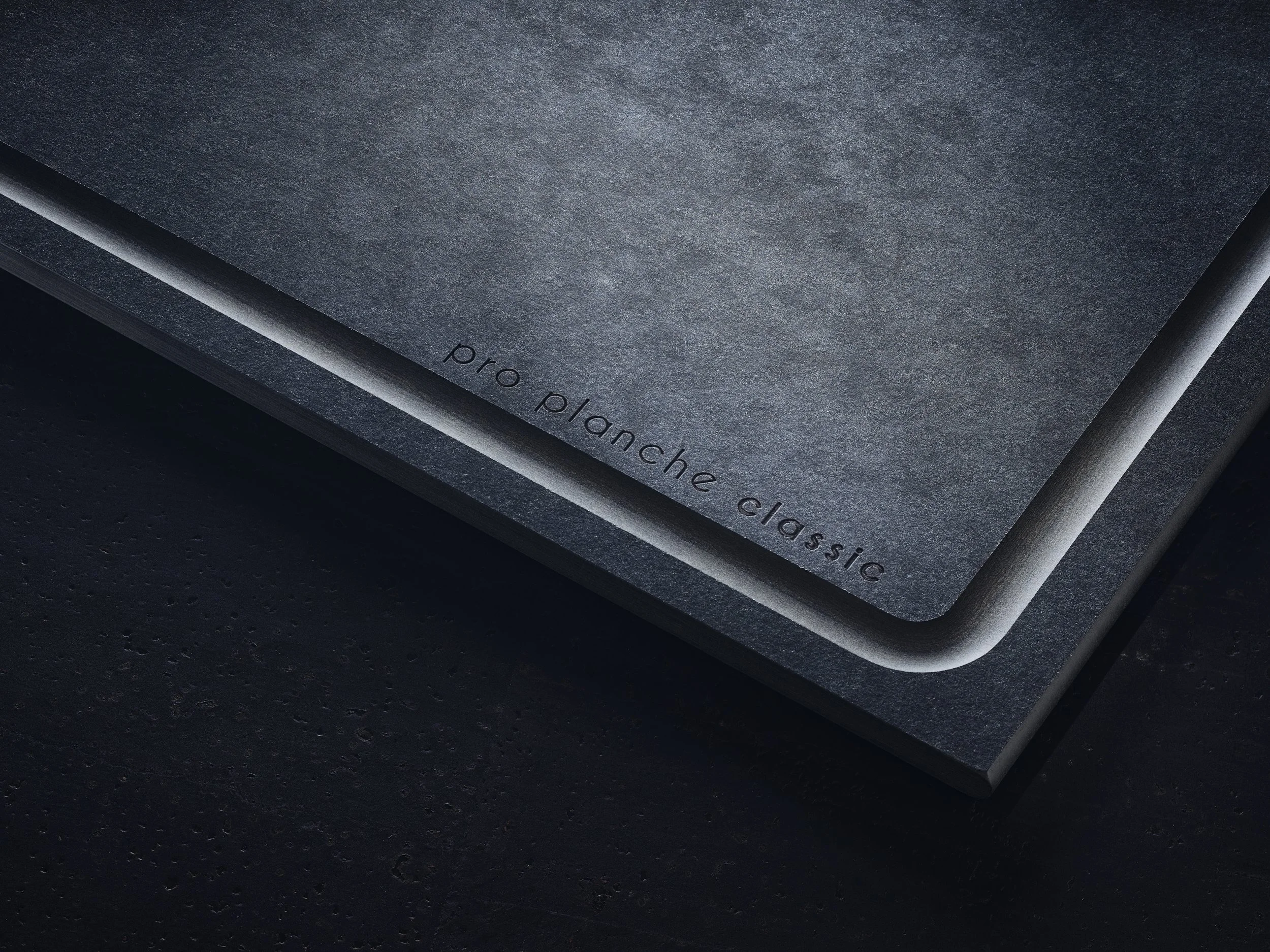 Nahaufnahme eines schwarzen Pro Planches Classic Brettes mit abgerundeten Ecken, auf dunklem Hintergrund, das Logo ist am unteren Rand sichtbar.