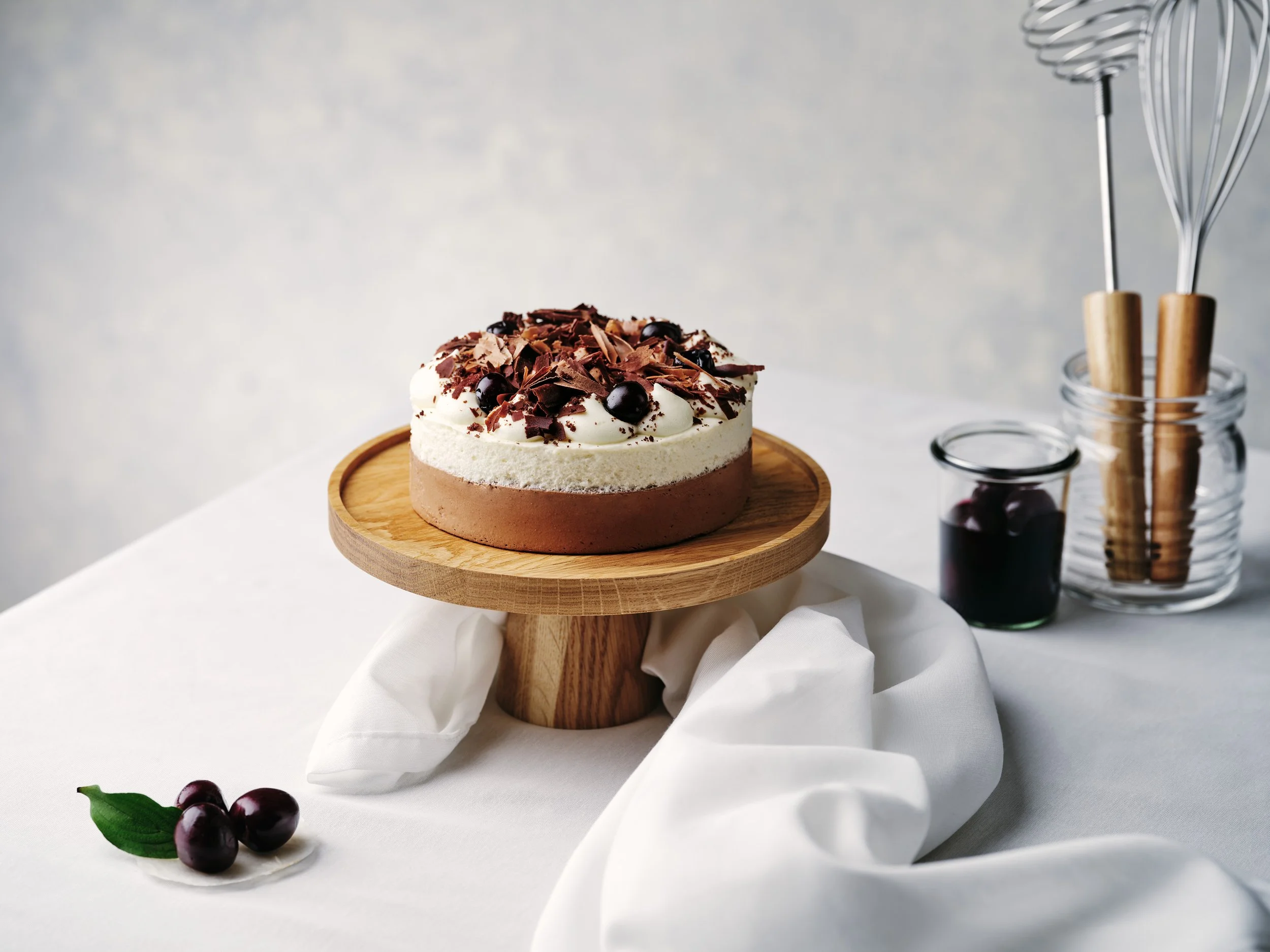 Schwarzwälder Kirschtorte auf einem hölzernen Tortenständer mit Schlagsahne, Schokoladenraspeln und Kirschen garniert. Im Hintergrund Glas mit Schneebesen und Glas mit Kirschen. Auf dem Tisch liegen Kirschen und ein Blatt.
