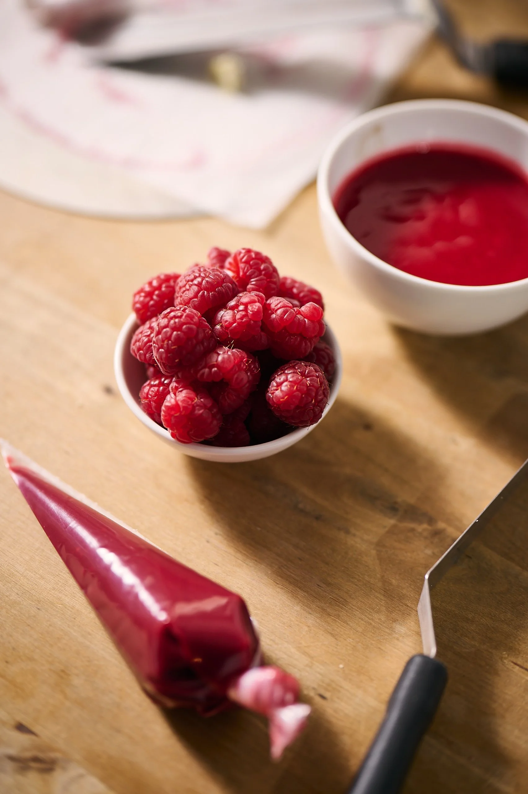 Nahaufnahme von Himbeeren in einer Schüssel auf einem Holztisch, im Hintergrund ist eine Tasse mit rotem Saft, auf dem Tisch liegen Küchenutensilien, vermutlich in einer Küche.