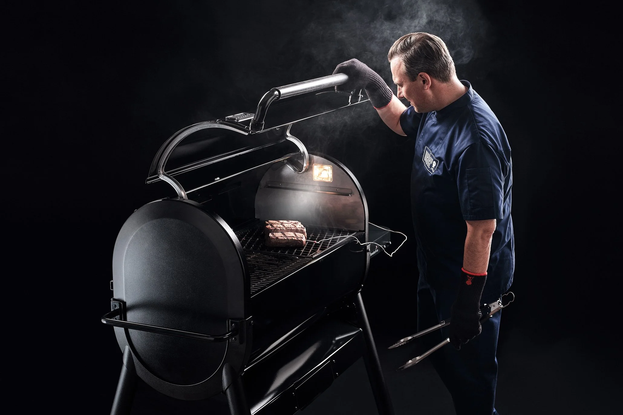 Mann grillt Fleisch in einem modernen Grill, schwarzer Hintergrund, Rauch steigt auf.